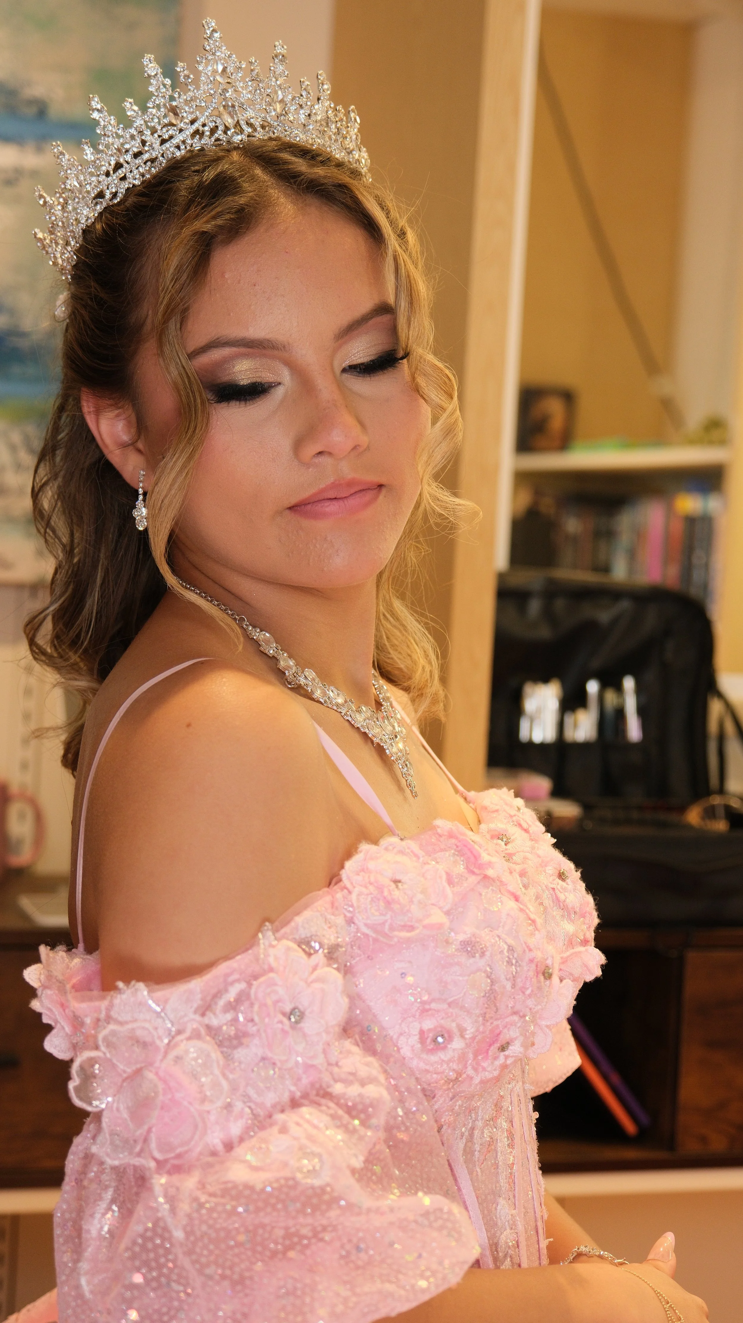 A young woman in a pink dress with floral embroidery, wearing a tiara and jewelry, appears to be at a formal event, possibly a pageant or celebration.