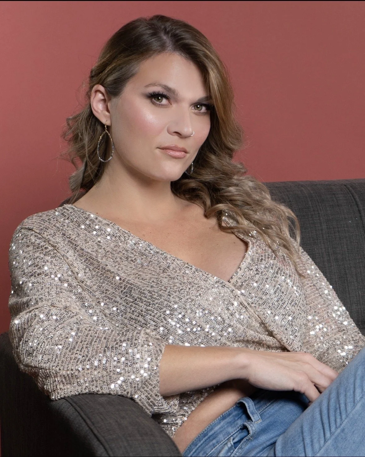 A young woman with wavy brown hair and hoop earrings sitting on a dark couch against a reddish wall, wearing a sparkly beige top and blue jeans.