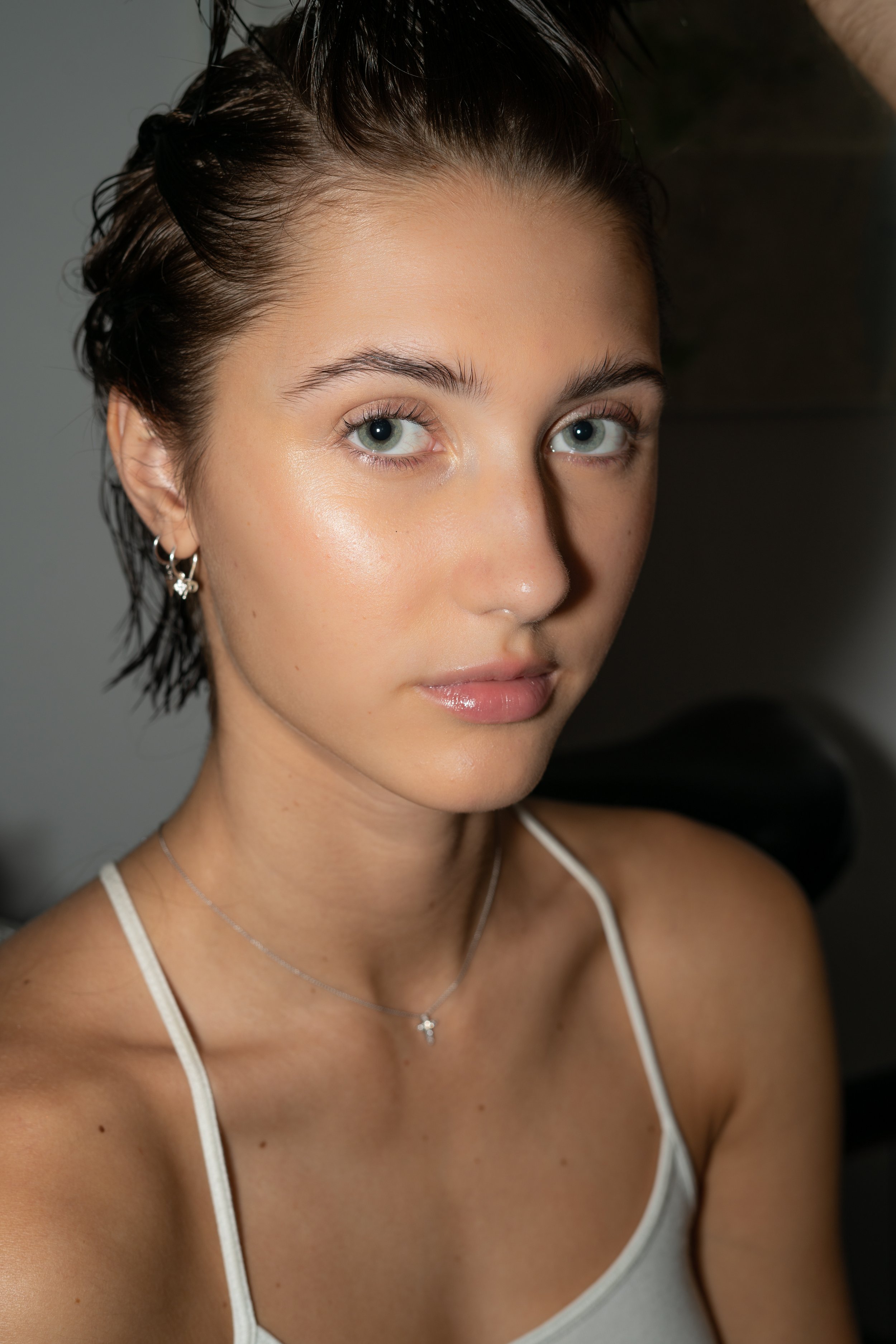 Close-up of a young woman with short, wet hair, wearing a white spaghetti strap top, earrings, and a silver necklace with a heart-shaped pendant. She has smooth, fair skin and striking blue eyes.