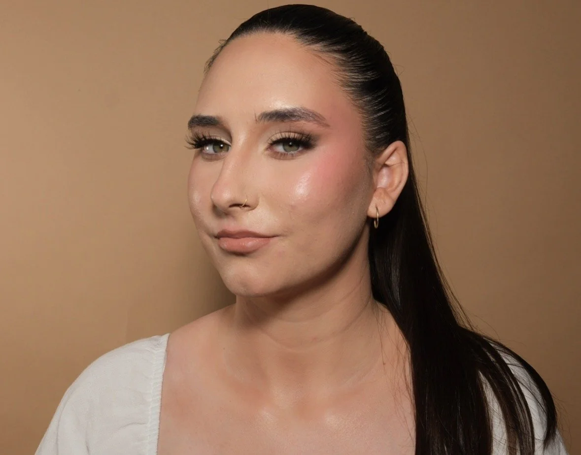 A woman with long dark hair pulled back, wearing makeup with bold eyebrows and earrings, posing against a beige background.