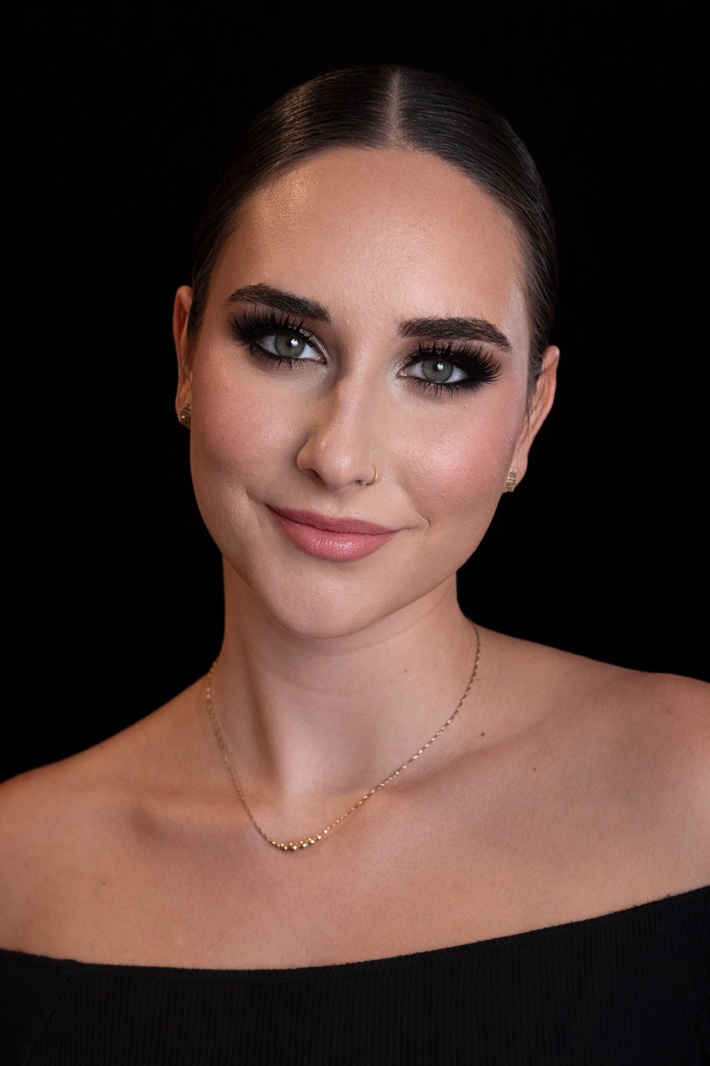 Close-up portrait of a young woman with dark hair pulled back, fair skin, bright eyes, and makeup, against a black background.