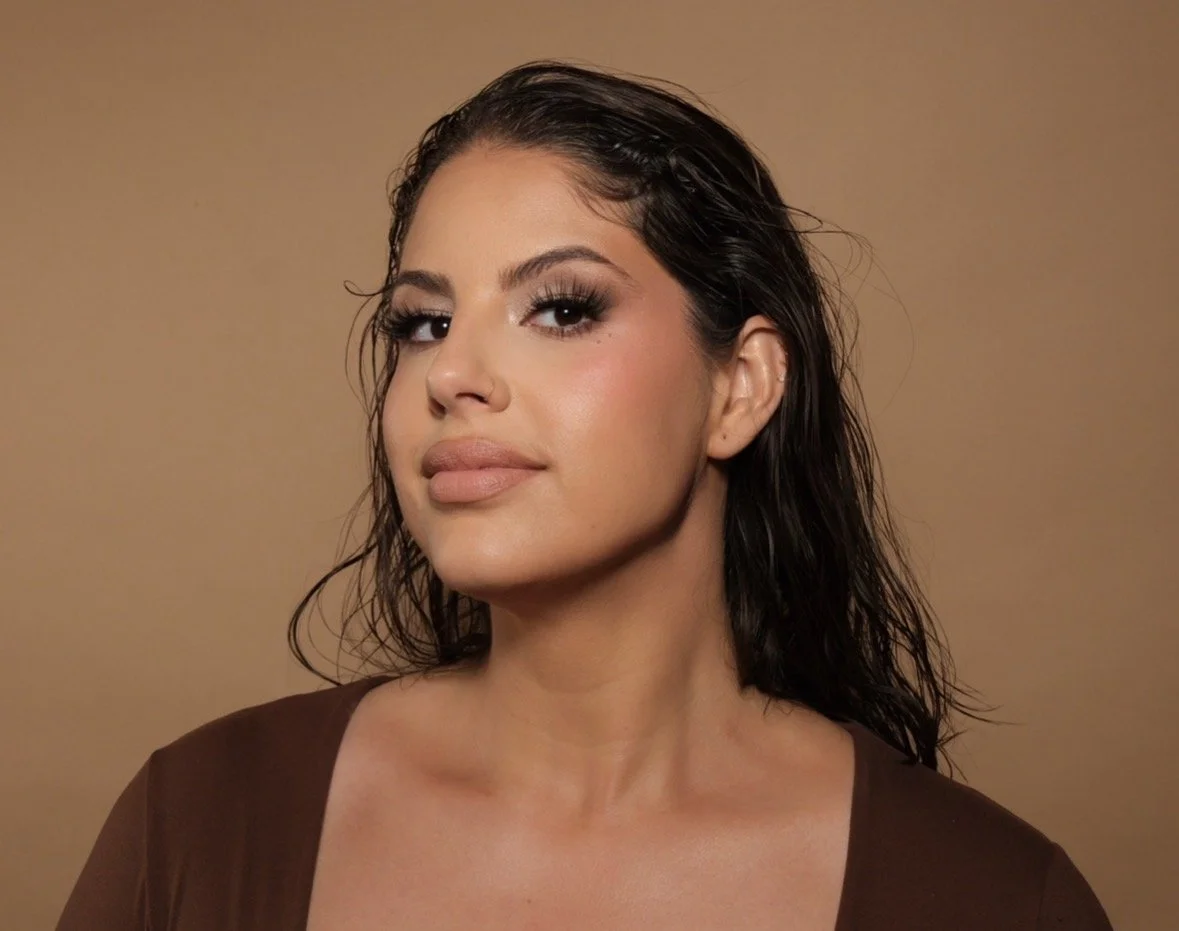 Close-up portrait of a woman with wet, dark hair, and makeup, wearing a brown top against a beige background.