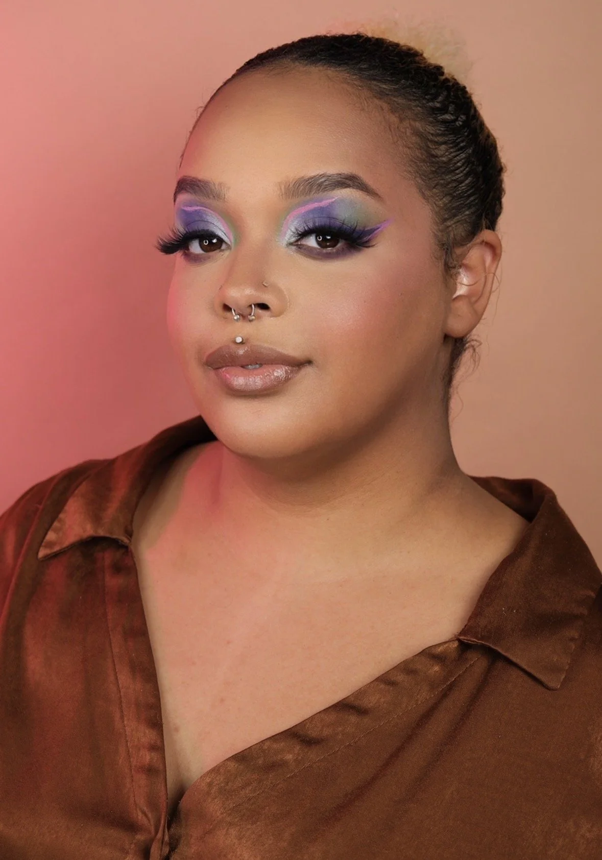 Close-up portrait of a woman with colorful makeup, including purple, green, and pink eyeshadow, long lashes, and nude lipstick; she has a septum piercing and short dark hair pulled back. She wears a brown satin shirt and stands against a pink backgro