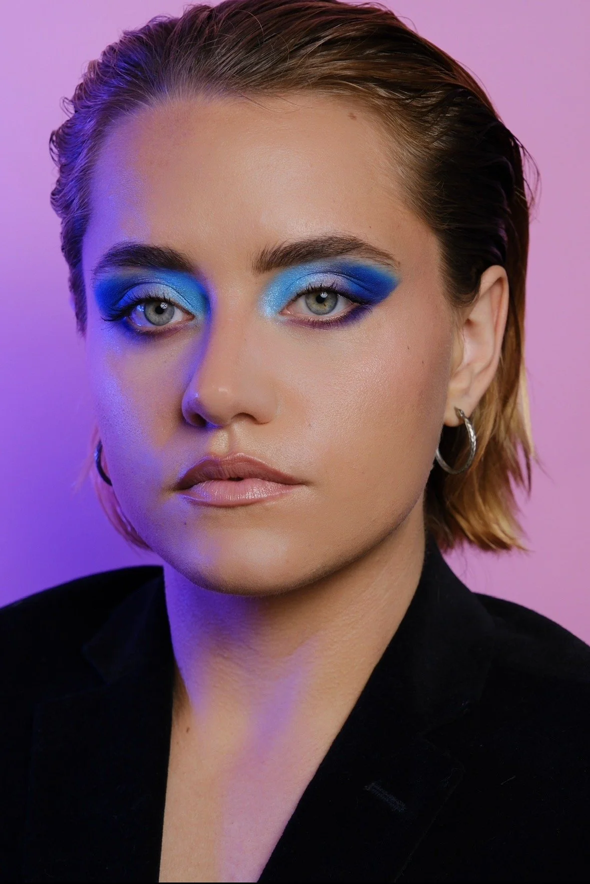 Close-up portrait of a woman with short, wet-looking hair and bold blue eyeshadow, wearing a black jacket and hoop earrings, against a soft purple background.