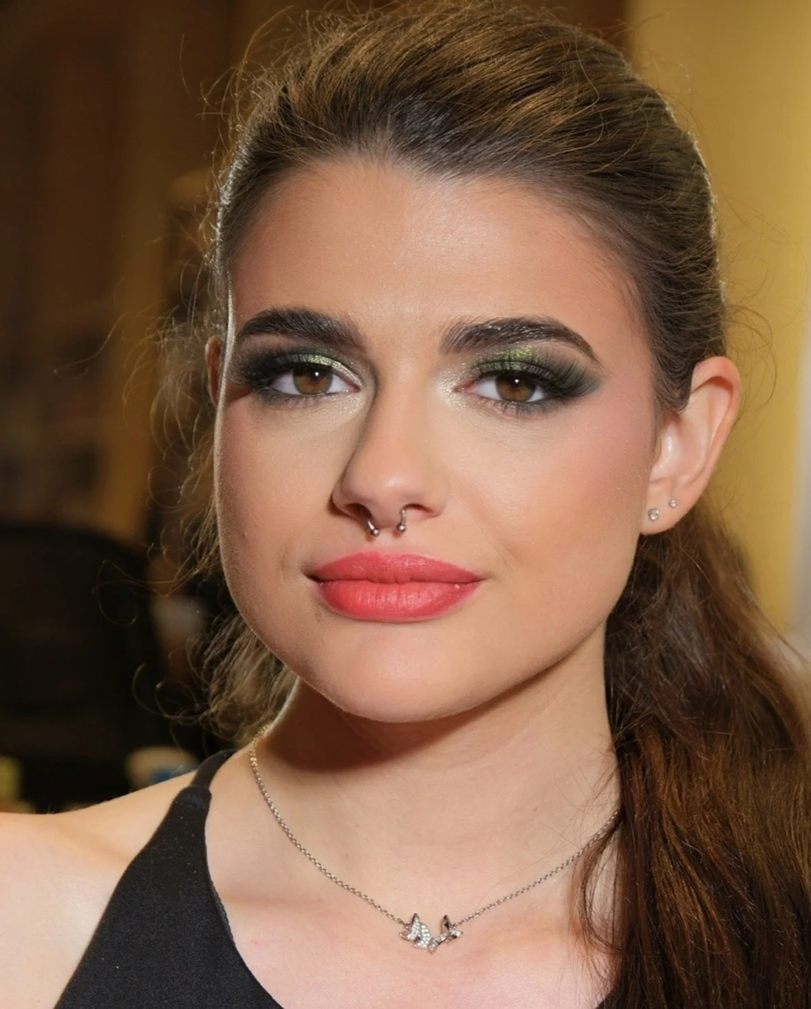 A woman with brown hair tied back, wearing makeup with bold eye shadow and pink lips, and sporting a septum piercing and small earrings. She wears a black top and a necklace with a butterfly pendant.