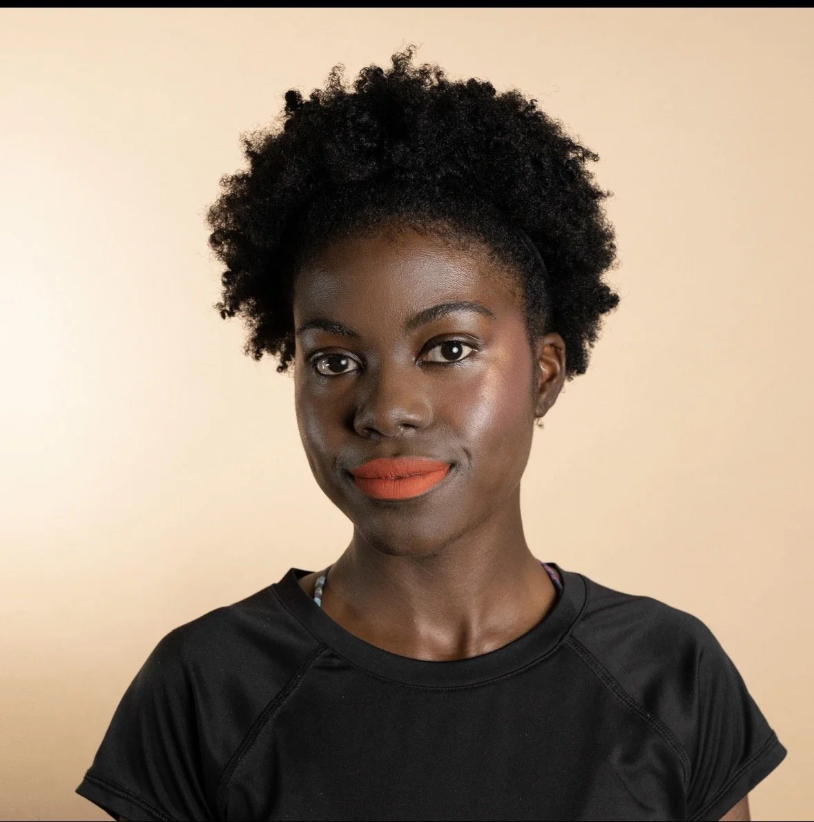 Portrait of a woman with natural black curly hair, wearing a black top, and orange lipstick against a beige background.