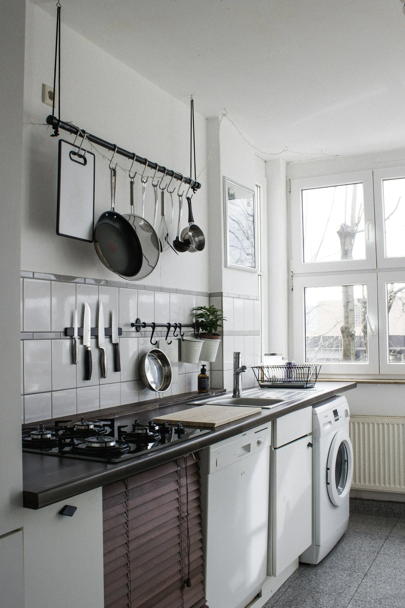 Kitchen with white cabinets, a gas stove with four burners, and a countertop with cleaning supplies. There is a rack above the counter holding various kitchen utensils including knives, pots, and a cutting board. A window lets in natural light, and a washing machine is under the counter.
