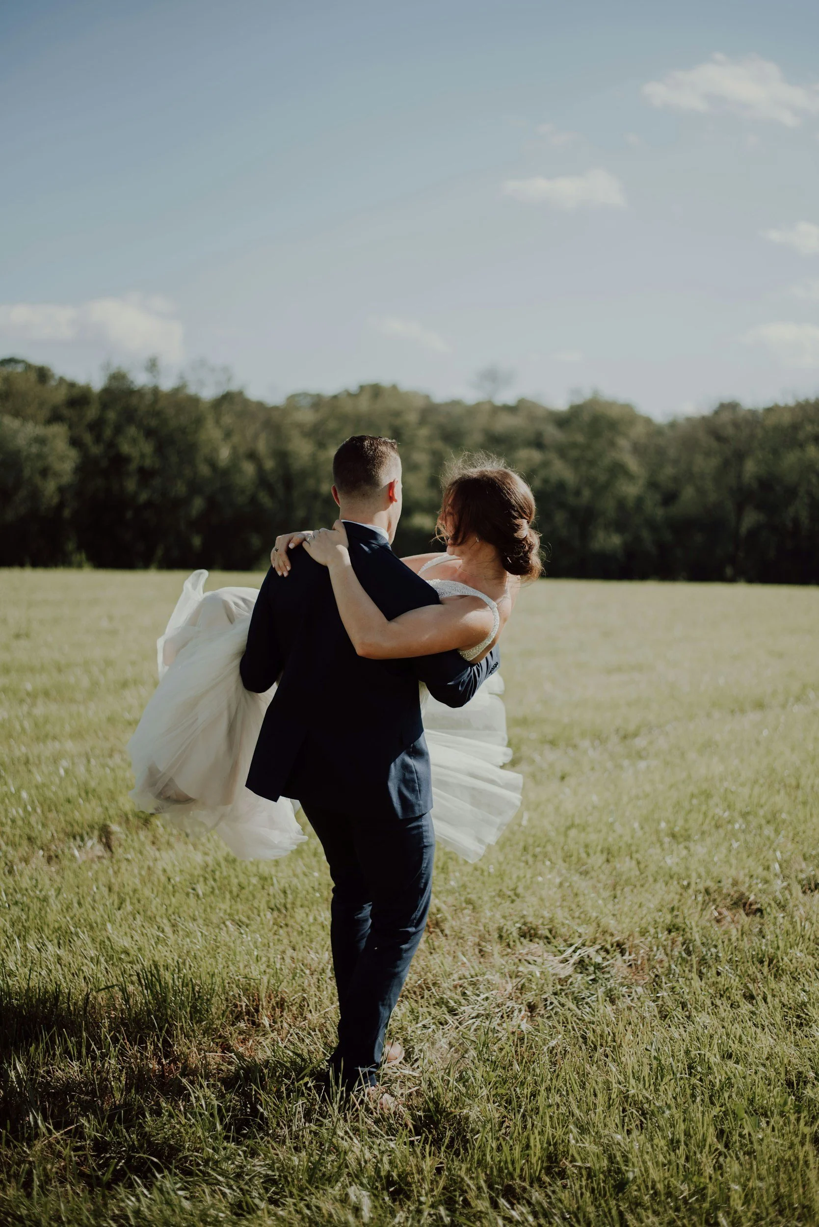 A groom carrying a bride in a white wedding dress across a grassy field outdoors on a sunny day.