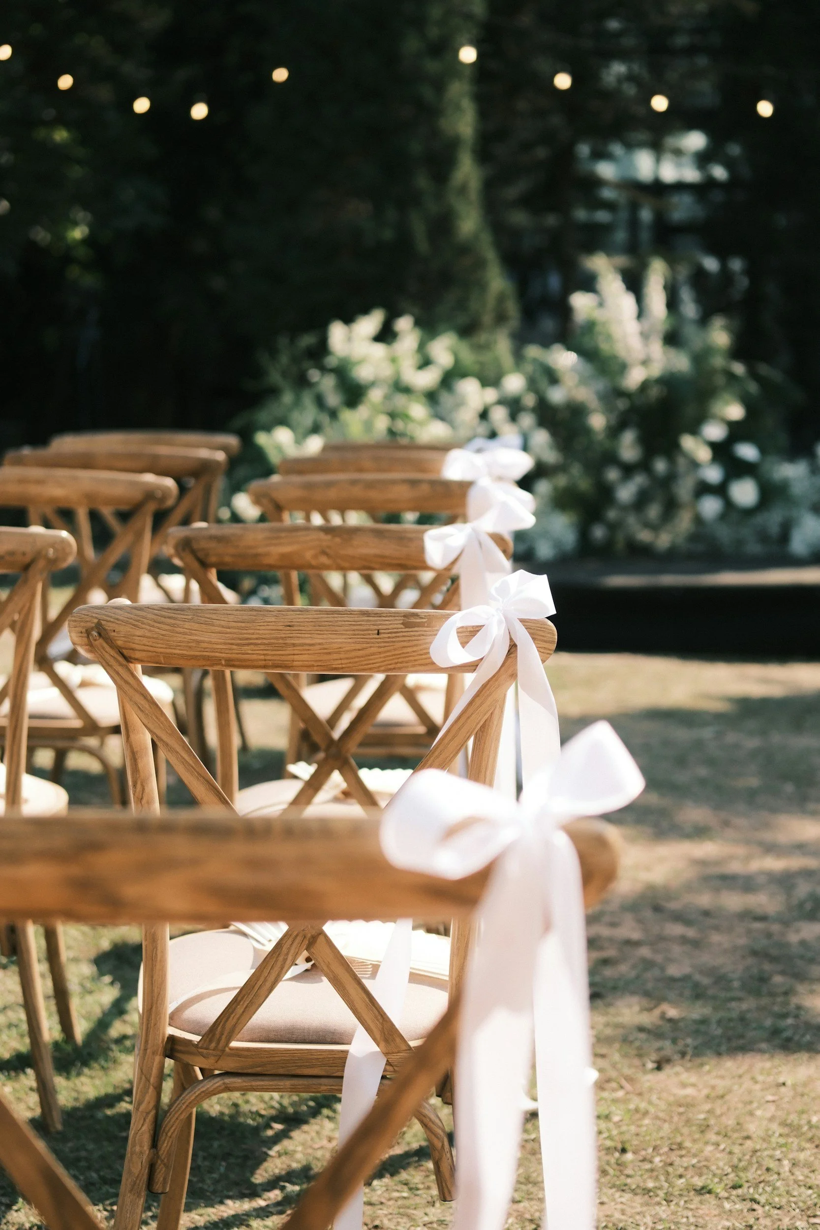 Wooden chairs with white ribbons tied on their backs, set outdoors, likely for a wedding or event, with greenery and string lights in the background.