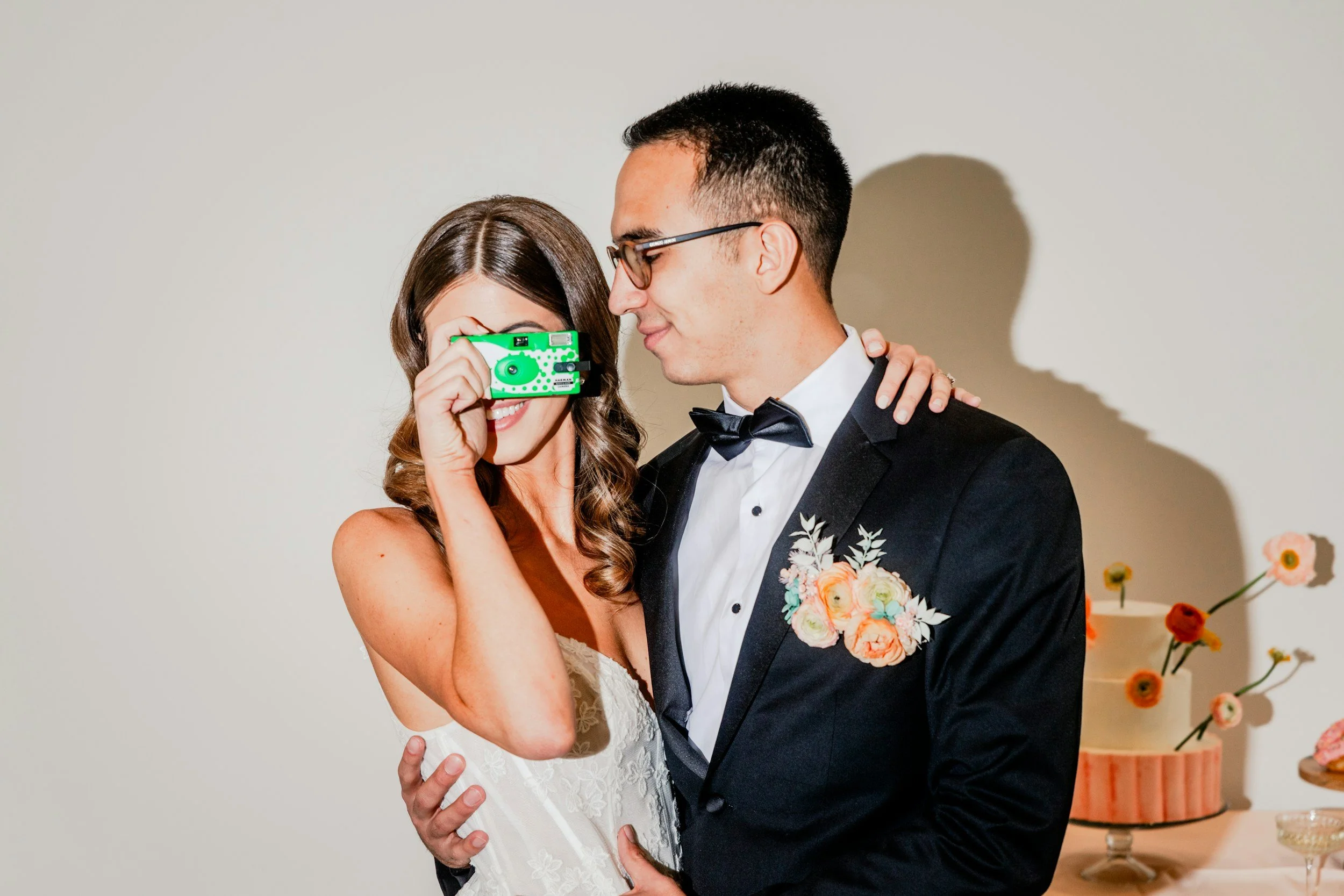A bride and groom are smiling and embracing each other at their wedding, with the bride holding a green camera up to her face.
