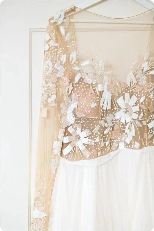 Close-up of a dress with floral and ribbon appliqué details, featuring sheer fabric with white and beige accents.