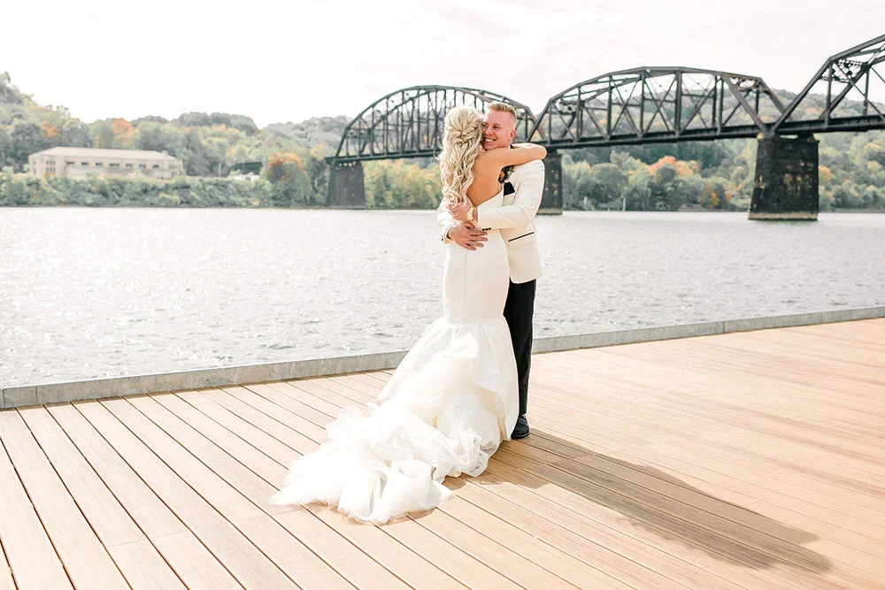 A bride and groom hugging on a wooden dock by a river with a bridge in the background, during daytime.