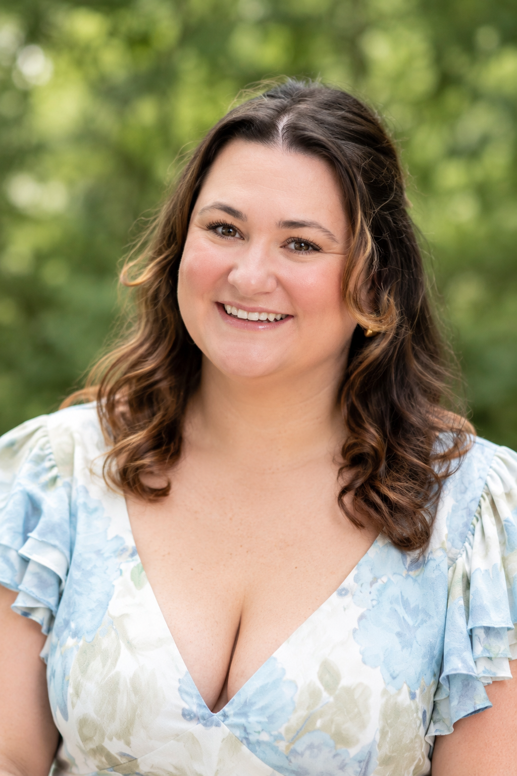 A smiling woman with wavy brown hair, wearing a light blue floral dress with ruffled sleeves, standing outdoors with green foliage in the background. Caitlyn Wimer