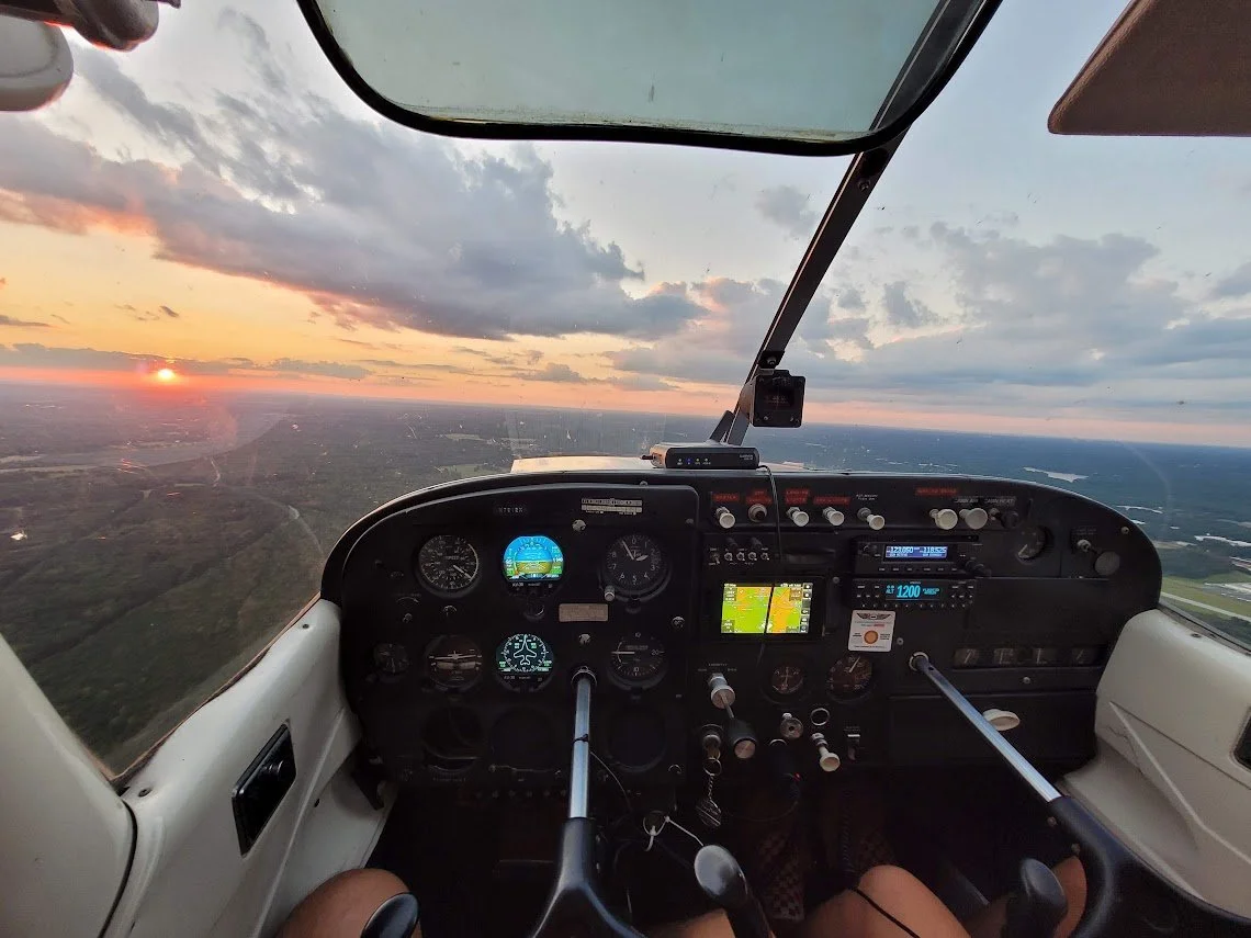 Inside view of a small airplane cockpit flying at sunset, showing the instrument panel and the pilot's legs.