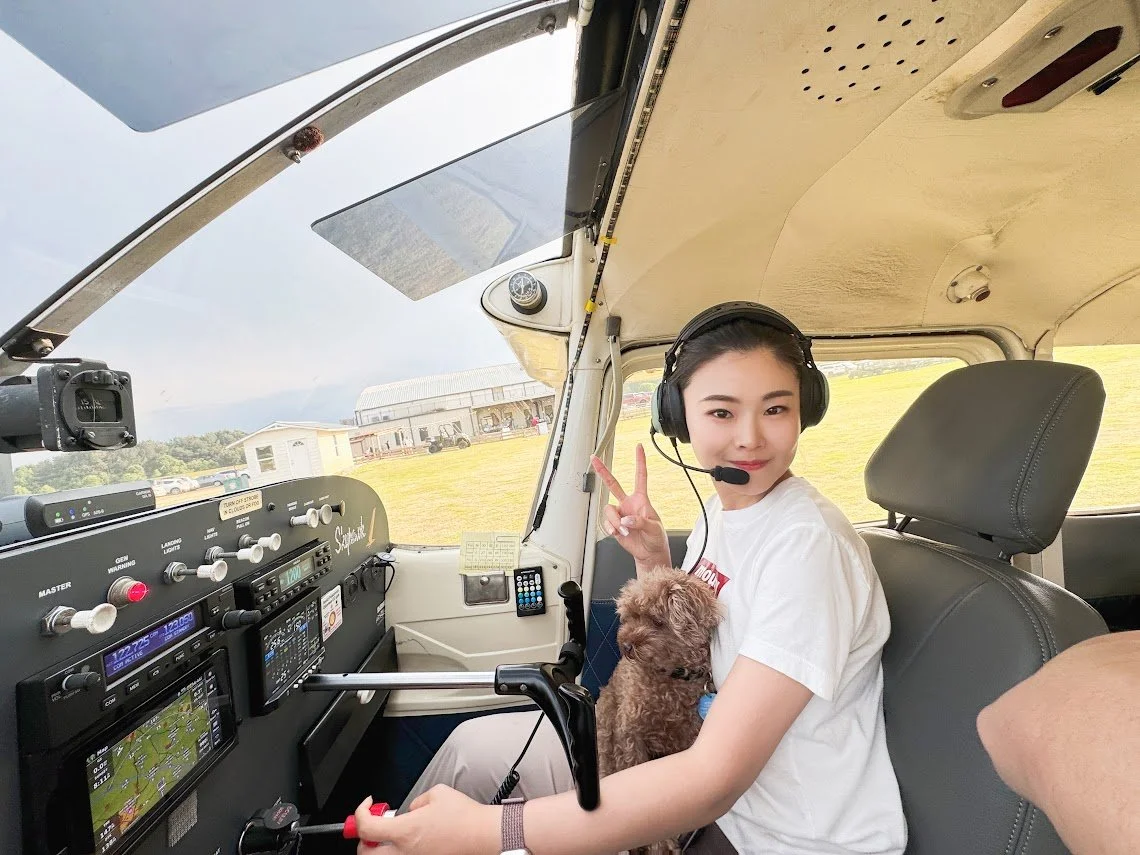 Young woman sitting in the cockpit of a small airplane, wearing a headset with microphone, making a peace sign with her fingers, holding a small dog, on a grassy airfield with hangar in the background.