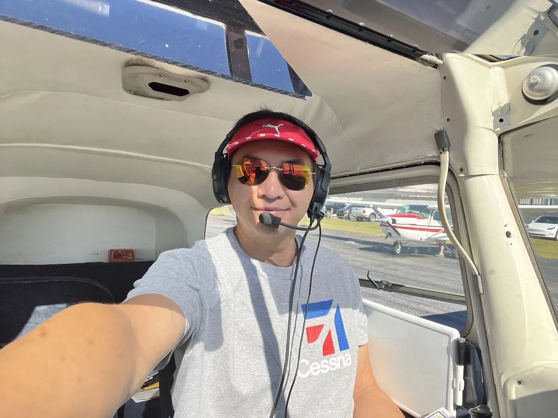 A man taking a selfie inside a small aircraft cockpit, wearing sunglasses, a red cap, a headset with microphone, and a gray t-shirt with a logo.
