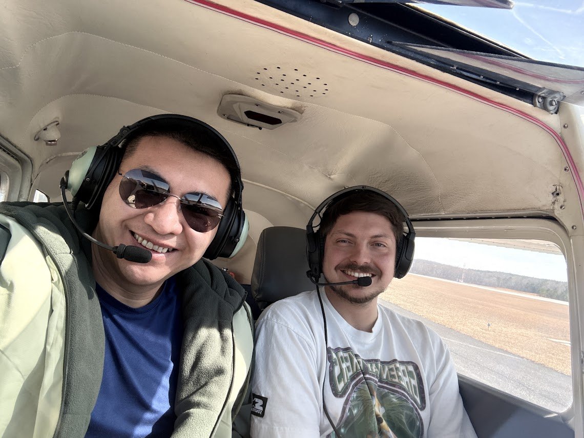 Two men sitting inside a small aircraft cockpit, wearing headsets and smiling, with open fields visible outside the window.