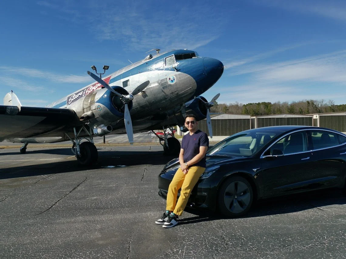 A man sitting on the front of a black Tesla, with a large vintage airplane in the background on an airport tarmac.