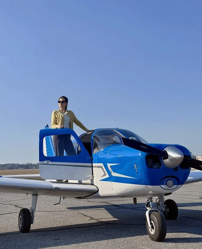 A woman in sunglasses and a yellow cardigan standing in the open cockpit of a blue and white small aircraft on an airfield.