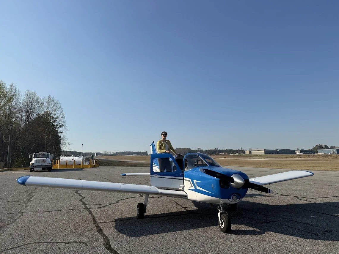 A person standing inside the open cockpit of a small blue and white propeller plane parked on an airport tarmac with a blue sky overhead.