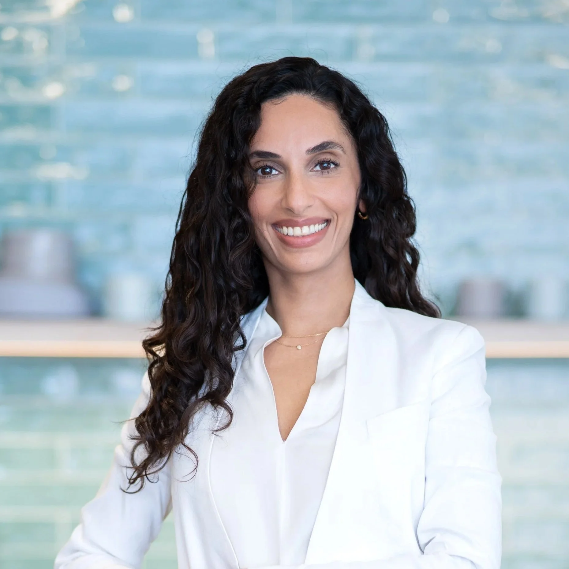 A woman with long, dark, curly hair smiling in front of a blurred indoor background with water-like bokeh lights, wearing a white shirt and a delicate necklace.