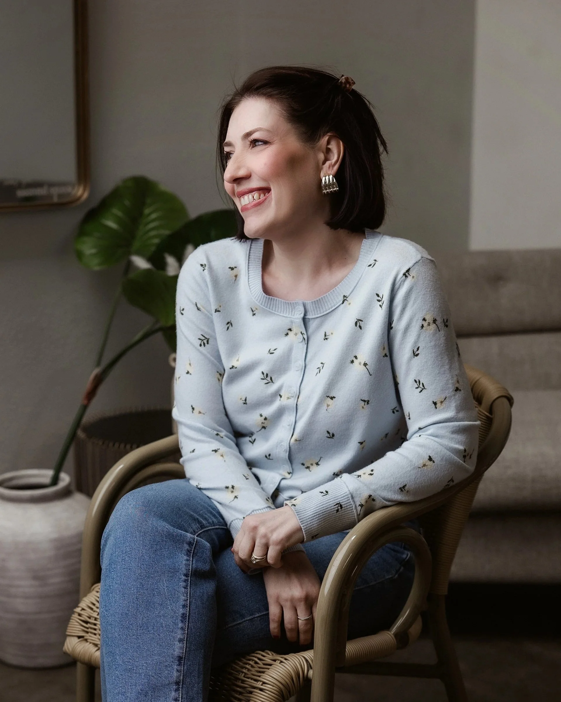 A woman sitting in a wicker chair with a smile on her face, wearing a light-colored sweater with a floral pattern, blue jeans, and silver earrings. There is a large green plant behind her and a wall with a mirror on the left.