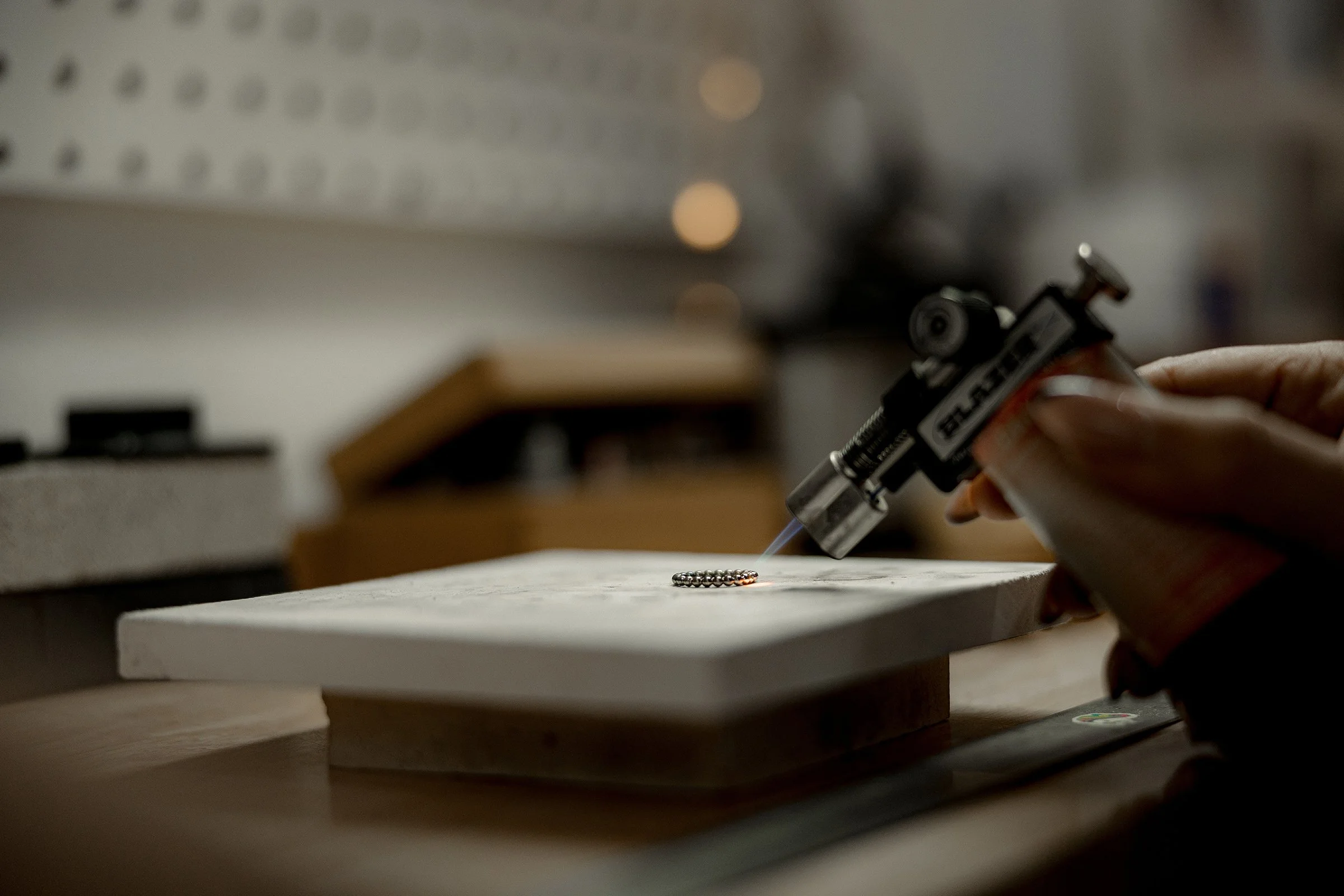 Person using a blowtorch to work on small metal beads on a work surface.