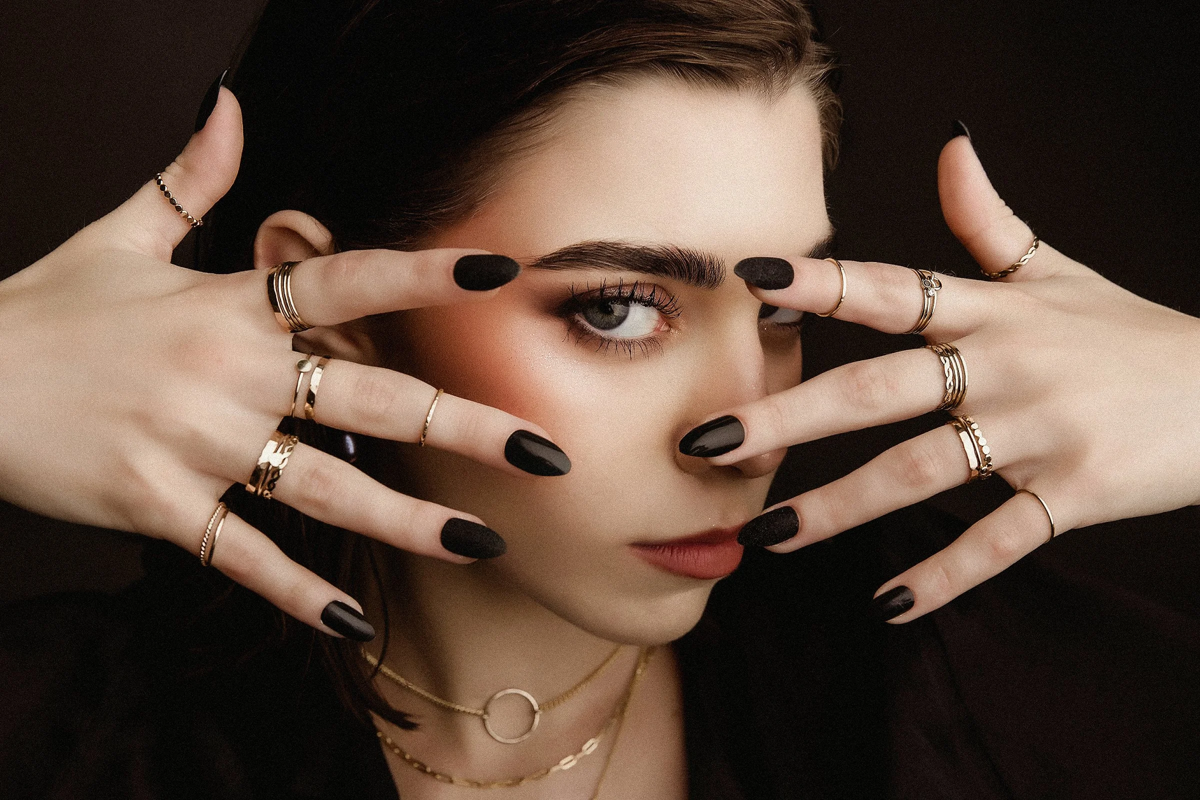 Close-up of a woman with dark hair and makeup, covering part of her face with both hands showing black nail polish and multiple rings and necklaces.