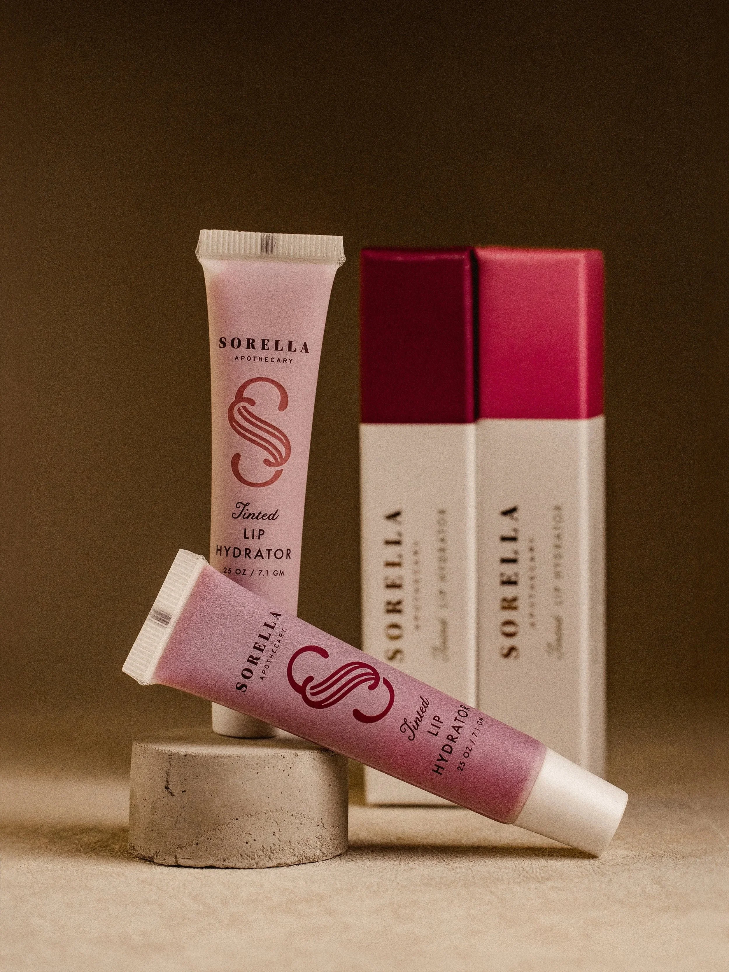 Pink tube labeled 'Sorella Apothecary Tinted Lip Hydrator' leaning against a white rounded block with a pink box in the background.
