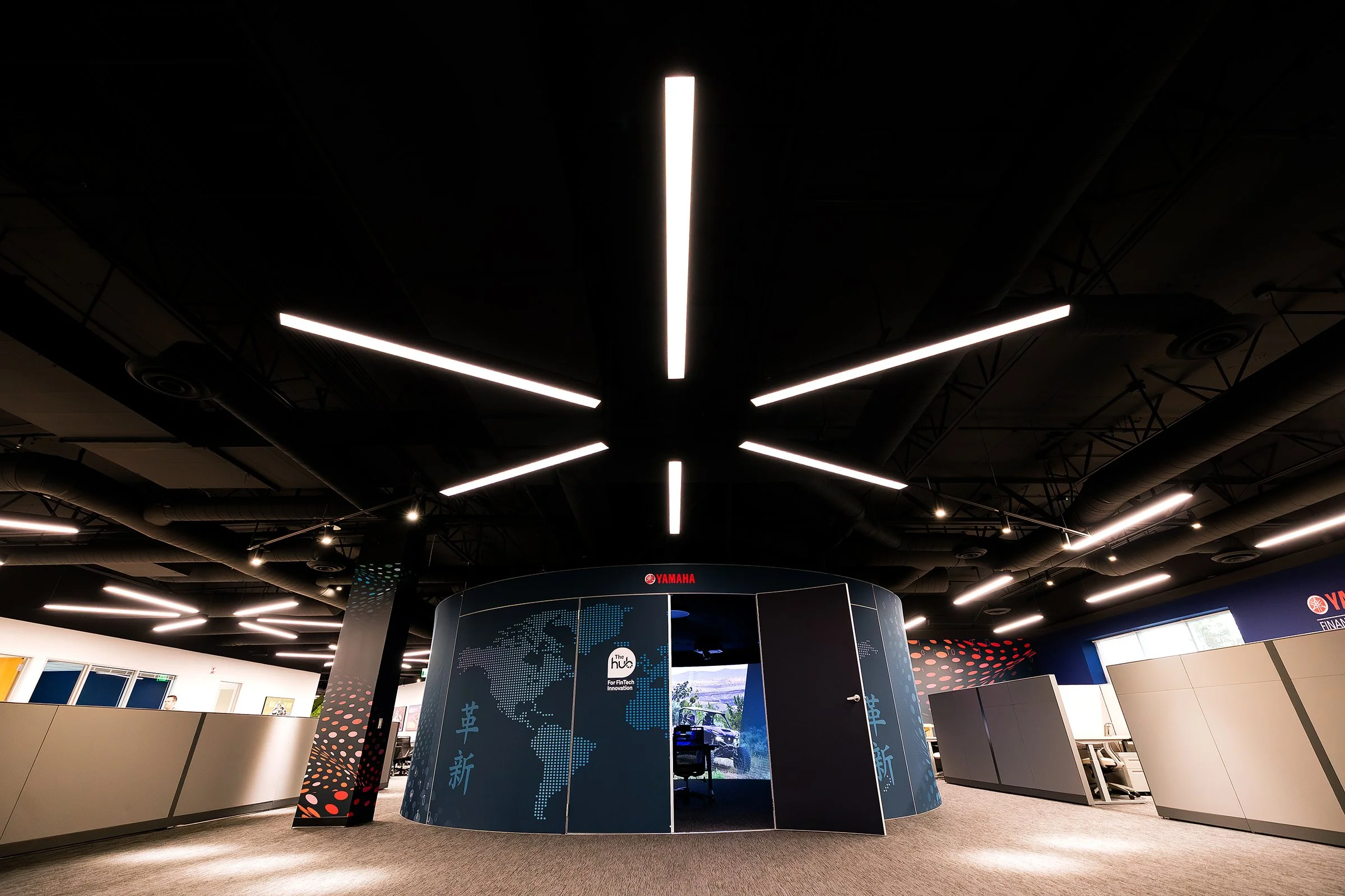 Interior of a modern office or exhibition space with a circular informational booth displaying a world map and Asian characters, surrounded by ceiling lights and industrial ductwork.