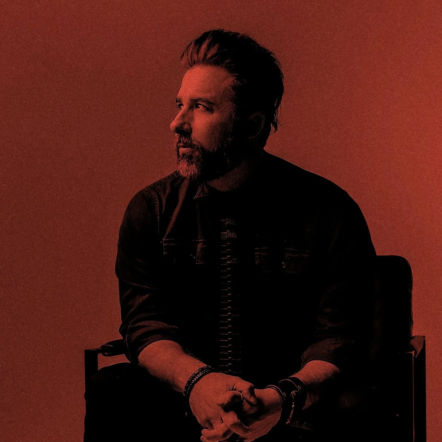 A man with a beard and styled hair, wearing a black jacket and wristbands, sitting against a red background, looking to the left.