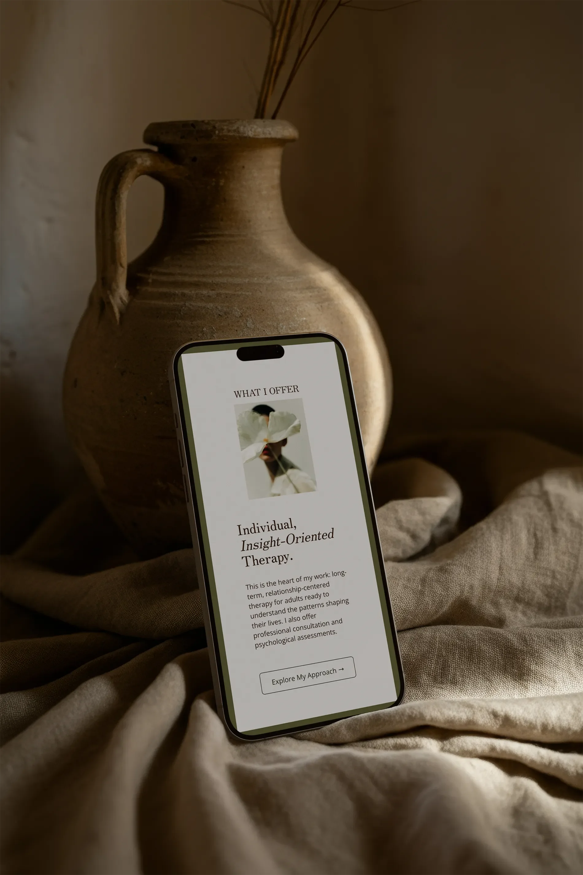 A smartphone displaying a webpage about therapy, placed on a beige fabric surface in front of a ceramic vase with dried grasses.