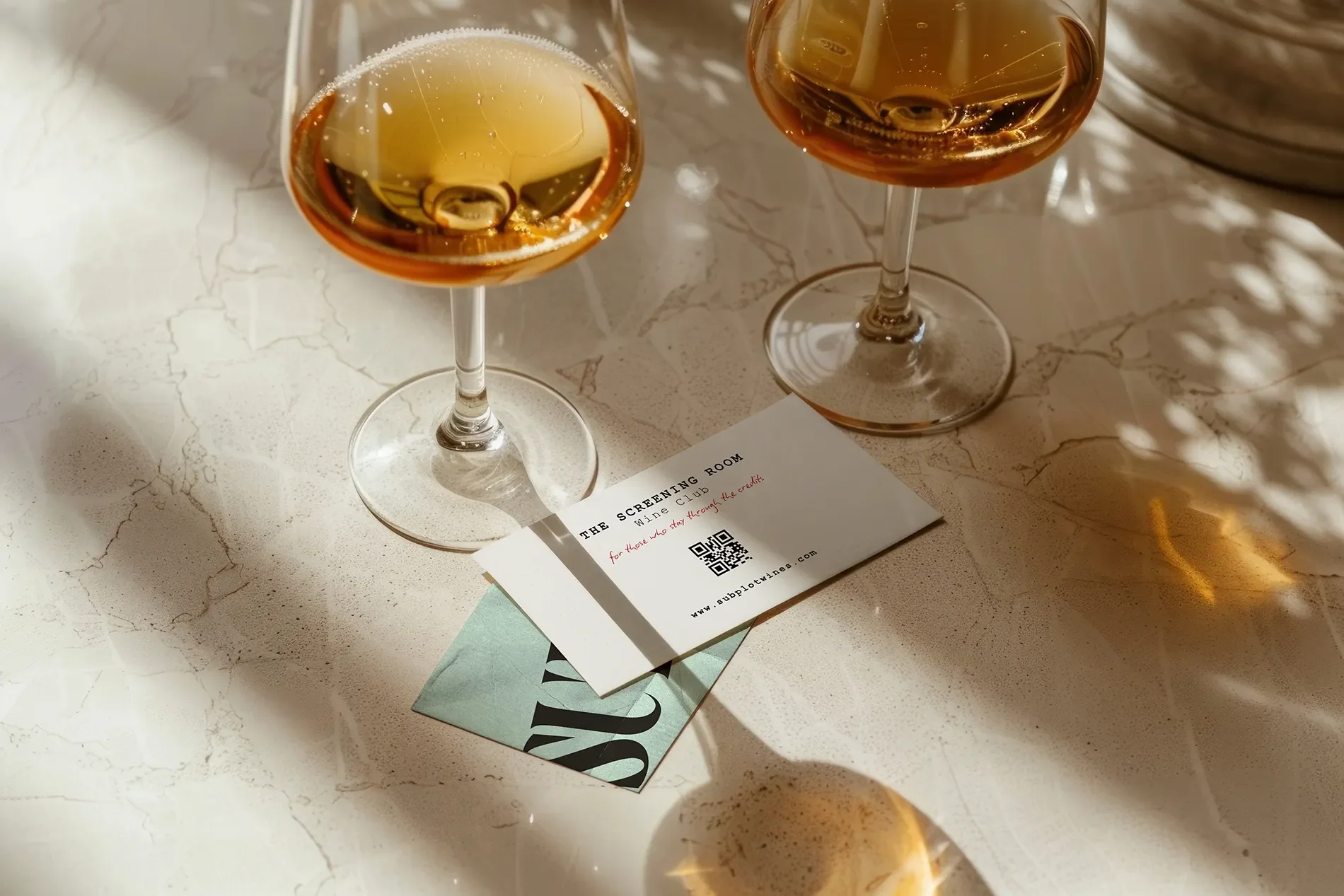 Two wine glasses with amber-colored wine on a white marble surface, alongside a printed card and a magazine.