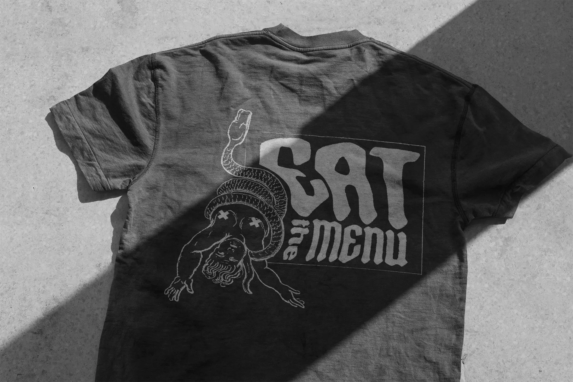 A black T-shirt with a graphic of a biblical figure, possibly a saint or a deity, with a snake wrapped around his head. The shirt has text that appears to read "SOT MISM" in a stylized font.