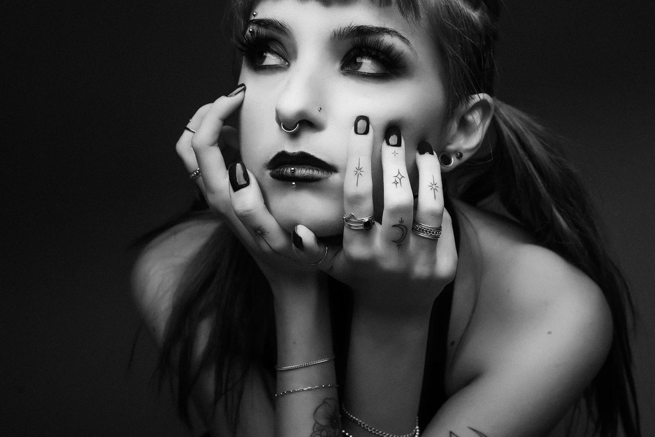 Black and white portrait of a woman with various facial piercings, tattoos on her fingers, and multiple rings and bracelets, touching her face with her fingers.
