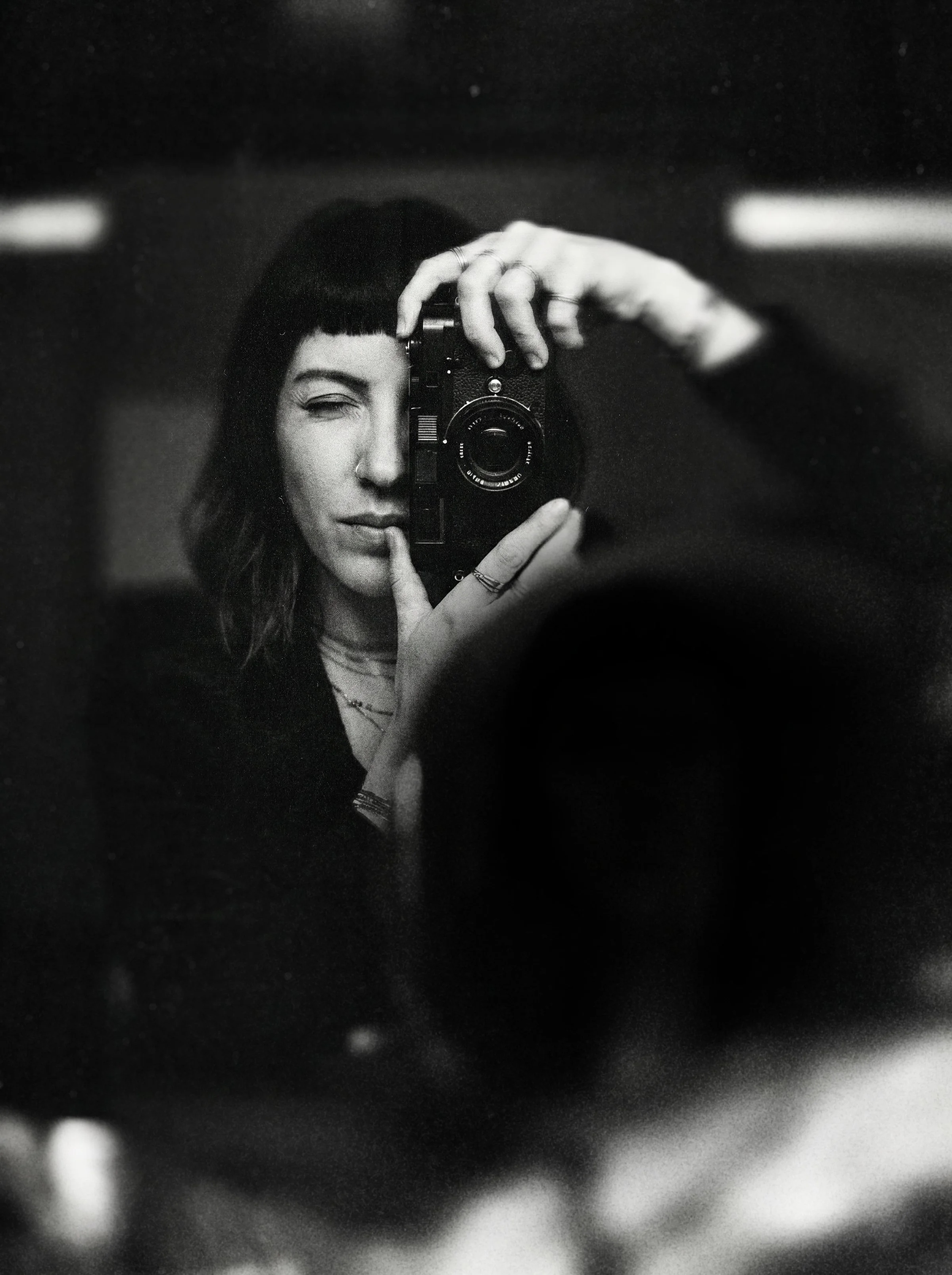 A woman taking a selfie in a mirror with a digital camera. She has dark hair, a nose ring, and is posing with one eye closed. The photo is in black and white.