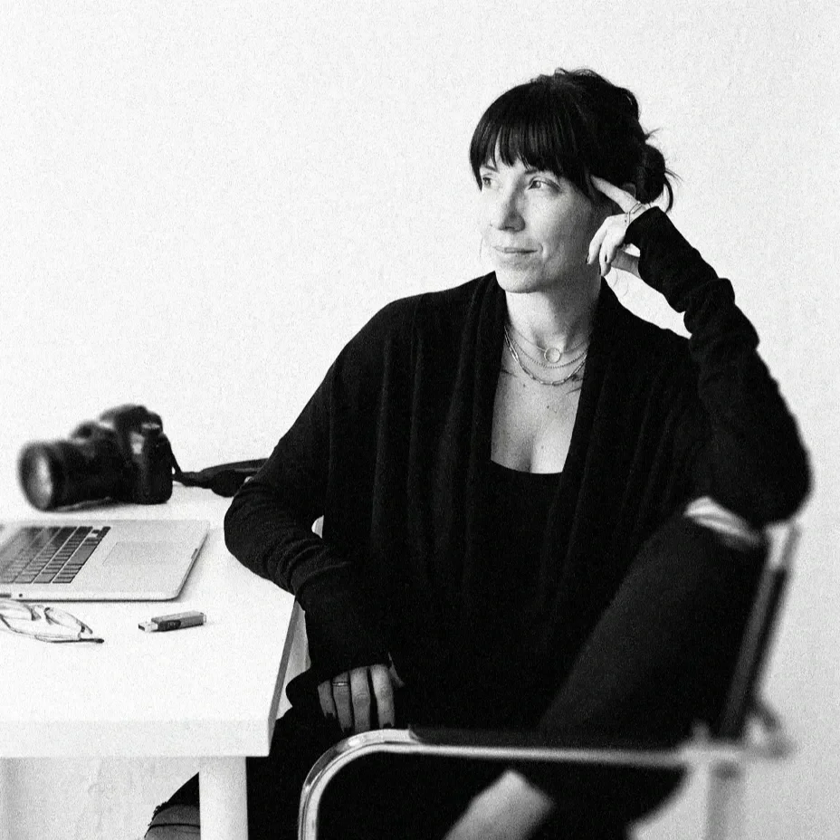 Black and white photo of a woman sitting at a desk with a laptop, camera, glasses, and a USB drive, resting her head on her hand, looking thoughtfully to the side.