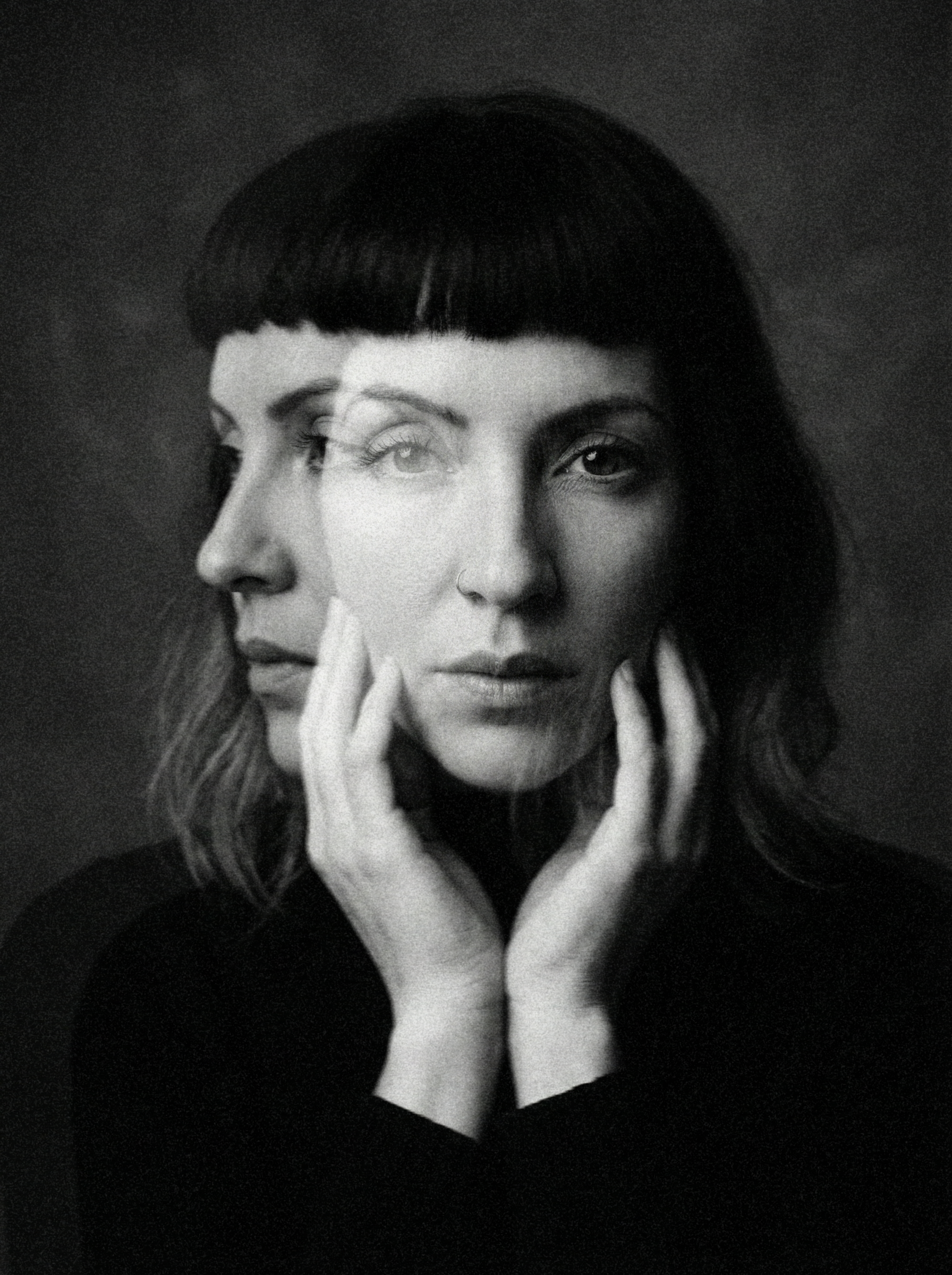 Black and white photograph of a woman with dark hair and a nose ring, posing with her fingers touching her face, looking directly at the camera.