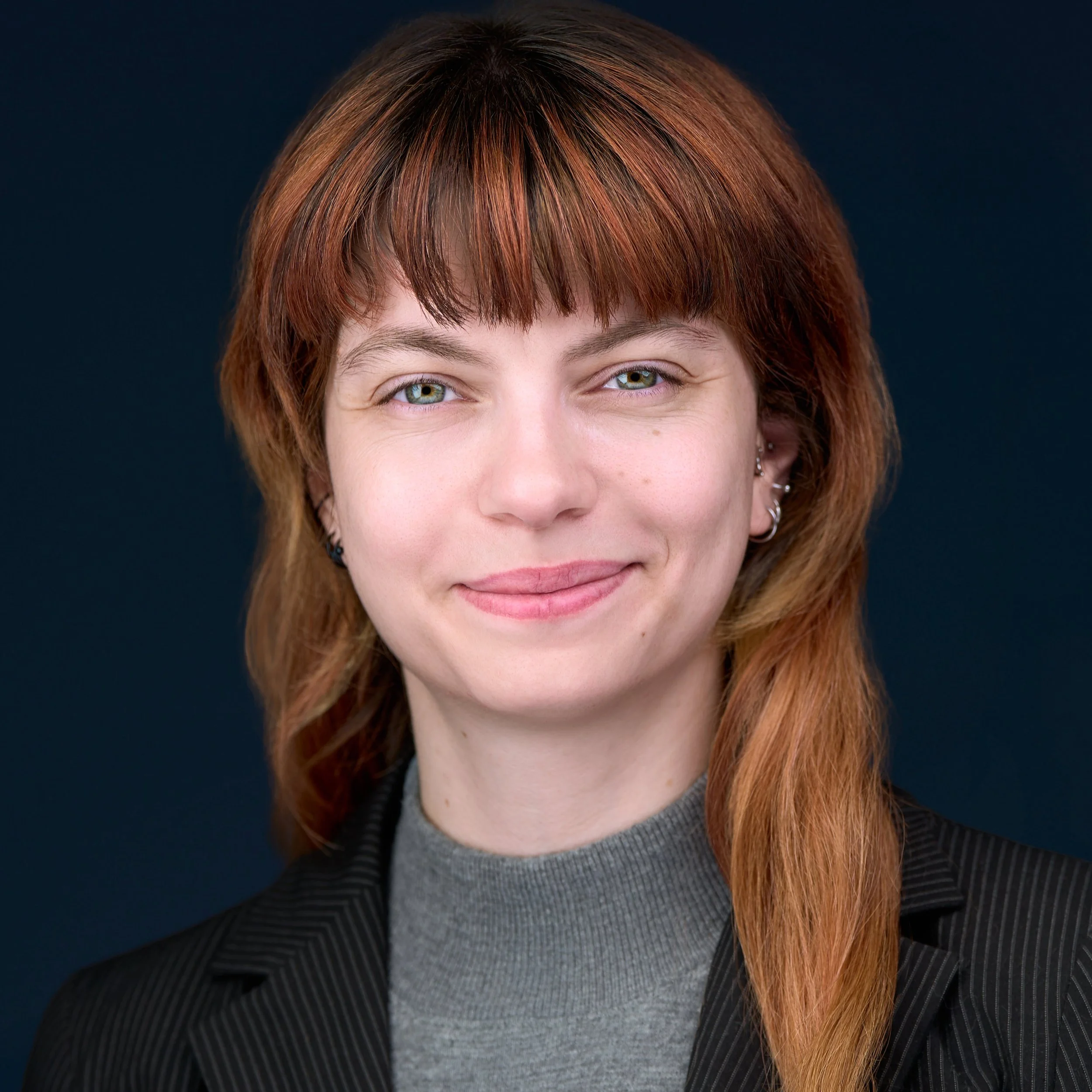 DAVIDSAGGIO Photography Tampa female executive portrait. Ginger hair with smile and direct eyes. Wearing gray top with black jacket against a very dark gray background.