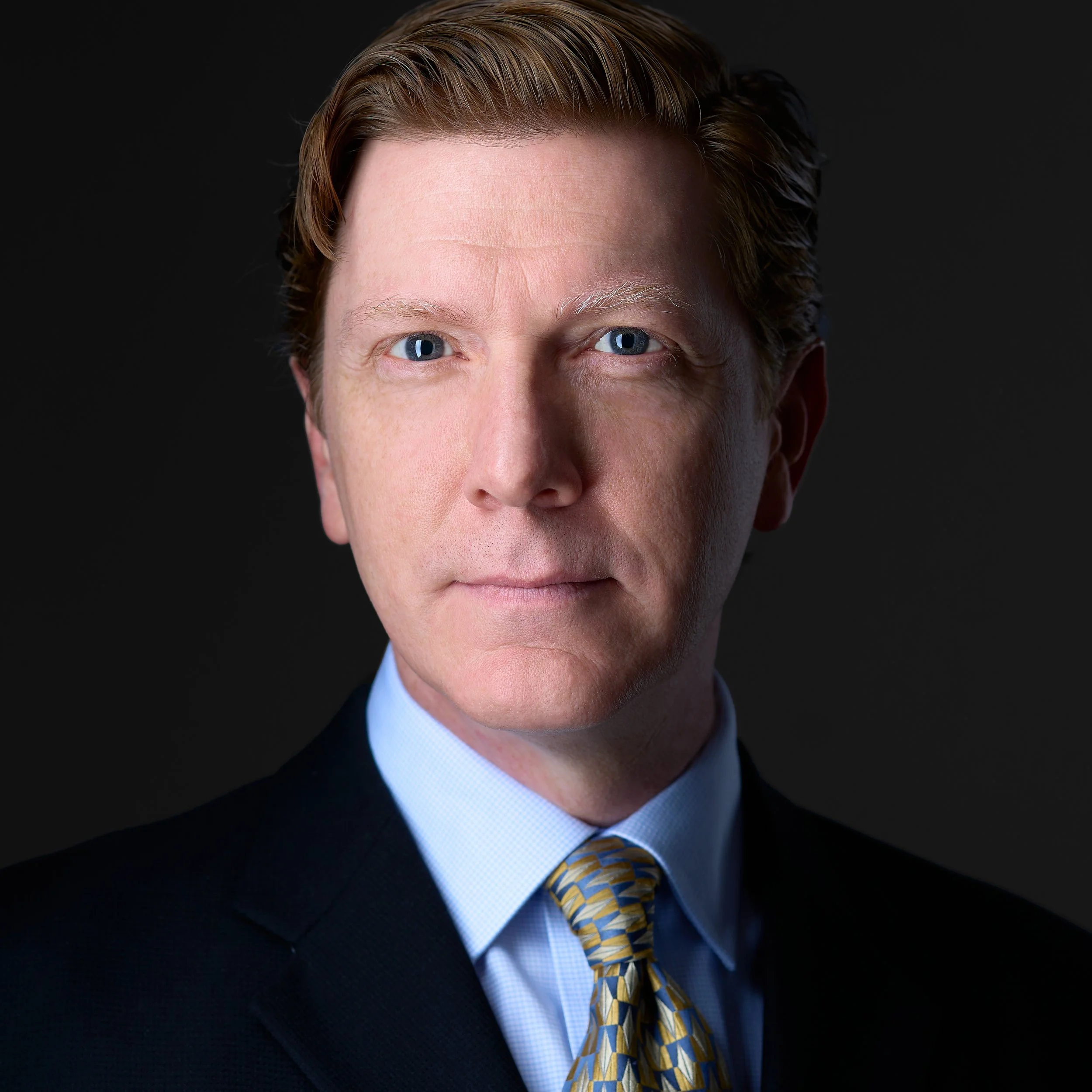 DAVIDSAGGIO Photography Tampa male executive portrait. Wearing a blue shirt, yellow patterned tie, and navy jacket. Rembrandt lighting.