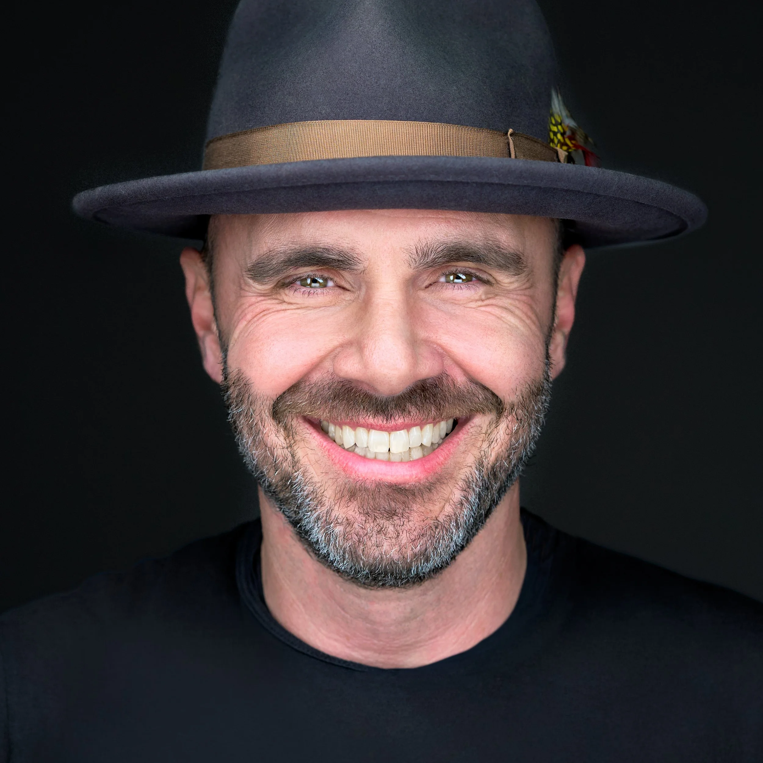 DAVIDSAGGIO Photography Tampa male entrepreneur headshot. Tightly cropped beard, large smile, and bright eyes. Wearing a fedora with feather and black crew neck top against a black background.