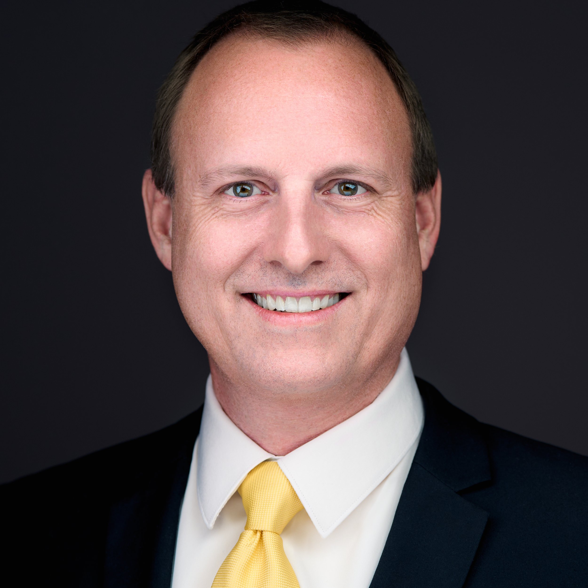 DAVIDSAGGIO Photography Tampa male financial executive headshot. Openly smiling. Wearing white shirt, yellow tie, and black jacket against dark gray background.