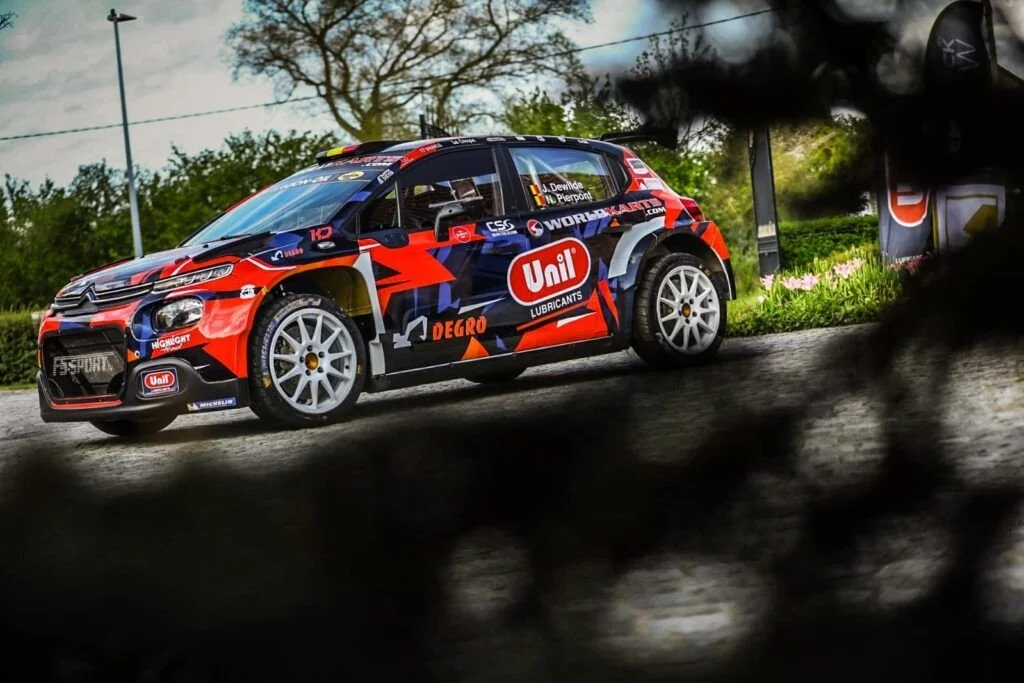 A rally car on a dirt road, partially obscured by a blurred foreground, with trees and grass in the background.
