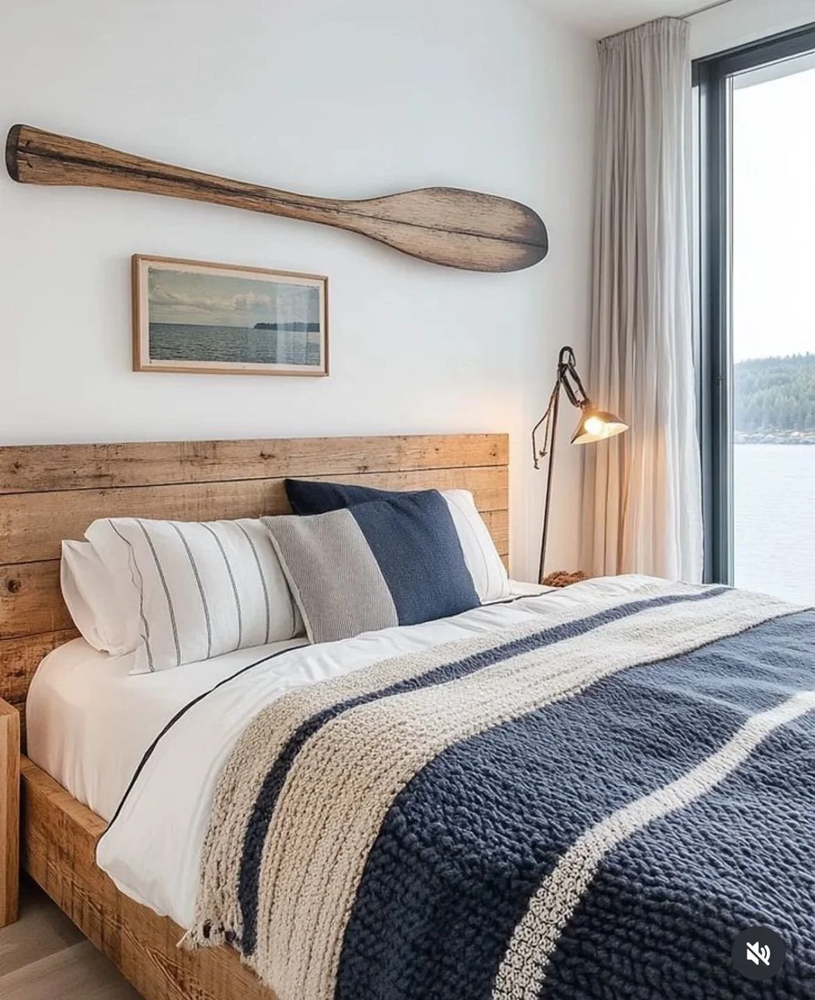 Ein Schlafzimmer mit einem grossen Bett, Kissen, einer gestrickten Decke, einem Holzkopfteil, einer Wandlampe, einer großen Fensterscheibe mit Blick auf Wasser und Natur, Wanddekoration in Form eines Paddels und einem Bild des Sees.