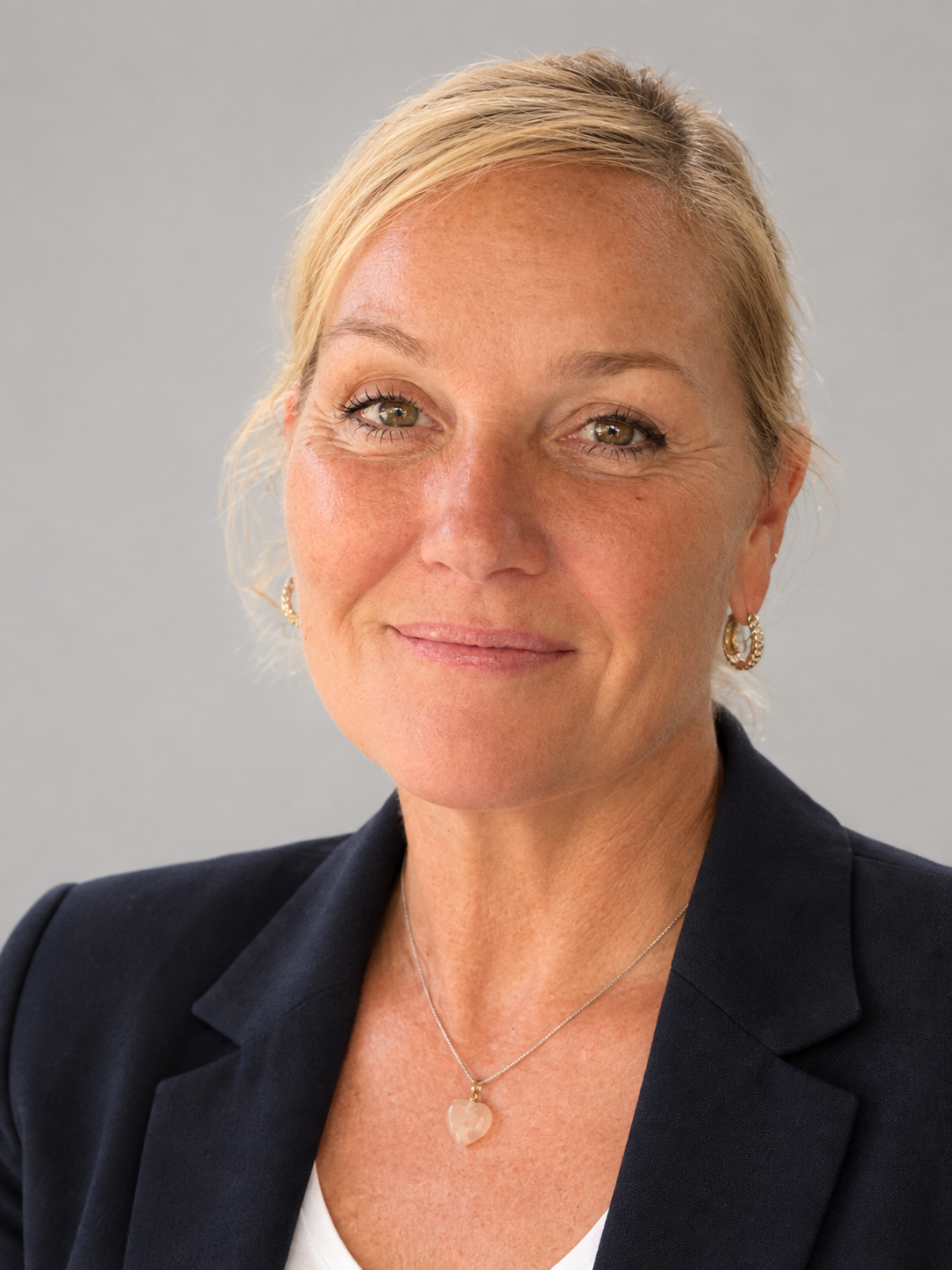 Nahaufnahme einer lachenden Frau mit blonden Haaren, goldenen Ohrringen, Kette mit Herzanhänger, schwarzem Blazer und weißem Top vor einem grauen Hintergrund.