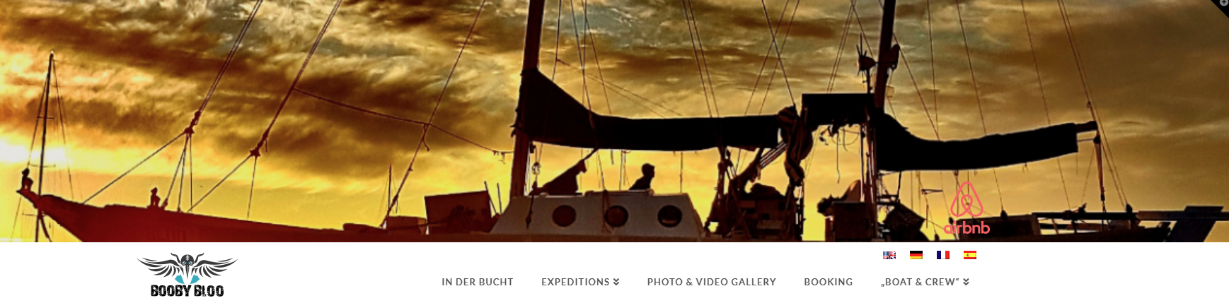 Sonnenuntergang über einem grossen Hochseekatamarant mit mehreren Personen an Bord, silhouettenhaft gegen den Himmel. Logo 'Booby Bloo' und Airbnb-Logo sichtbar.