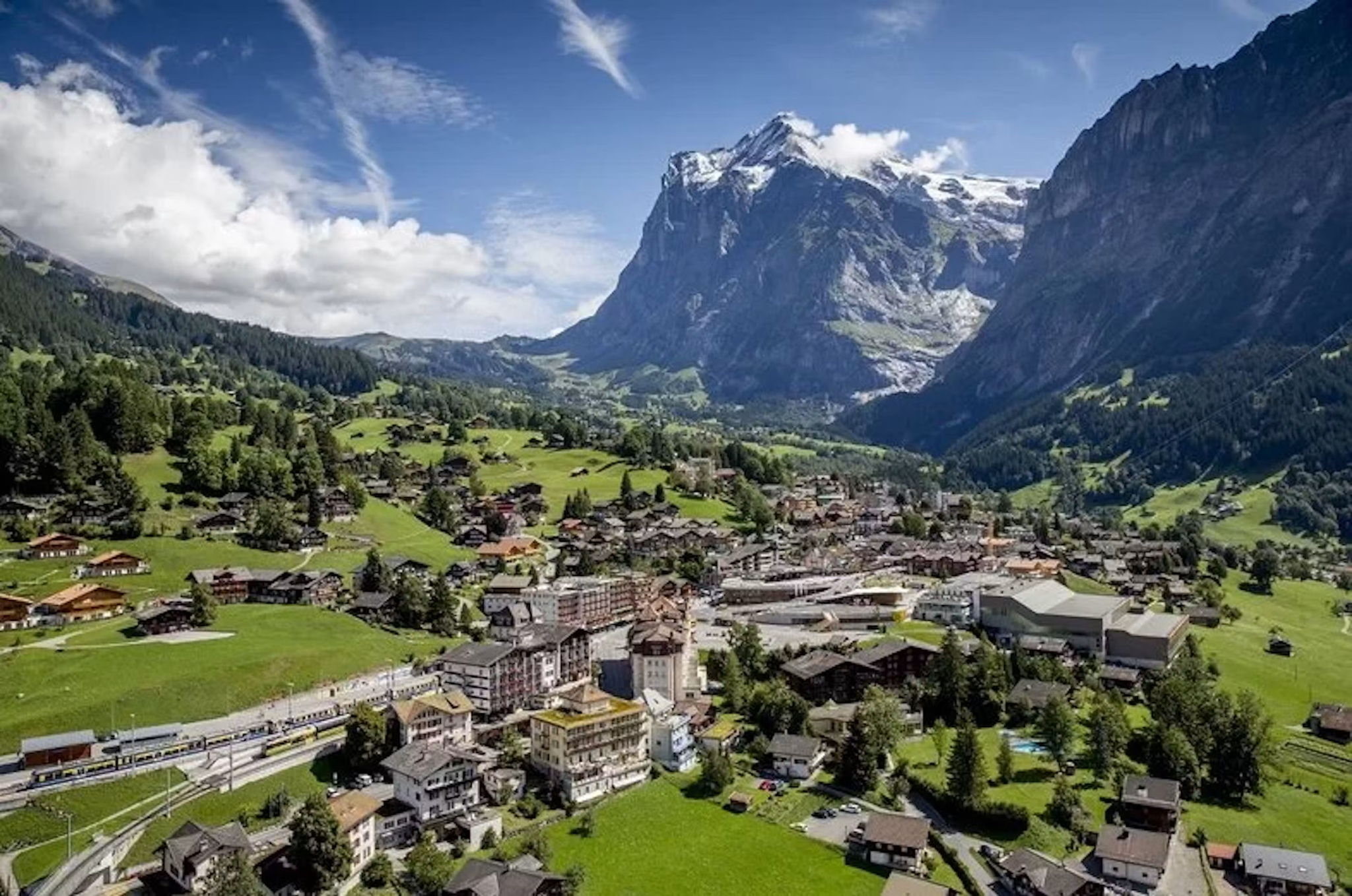 Eine malerische Schweizer Alpenlandschaft mit einem kleinen Dorf, grünen Wiesen und hohen, schneebedeckten Bergen im Hintergrund bei sonnigem Wetter.