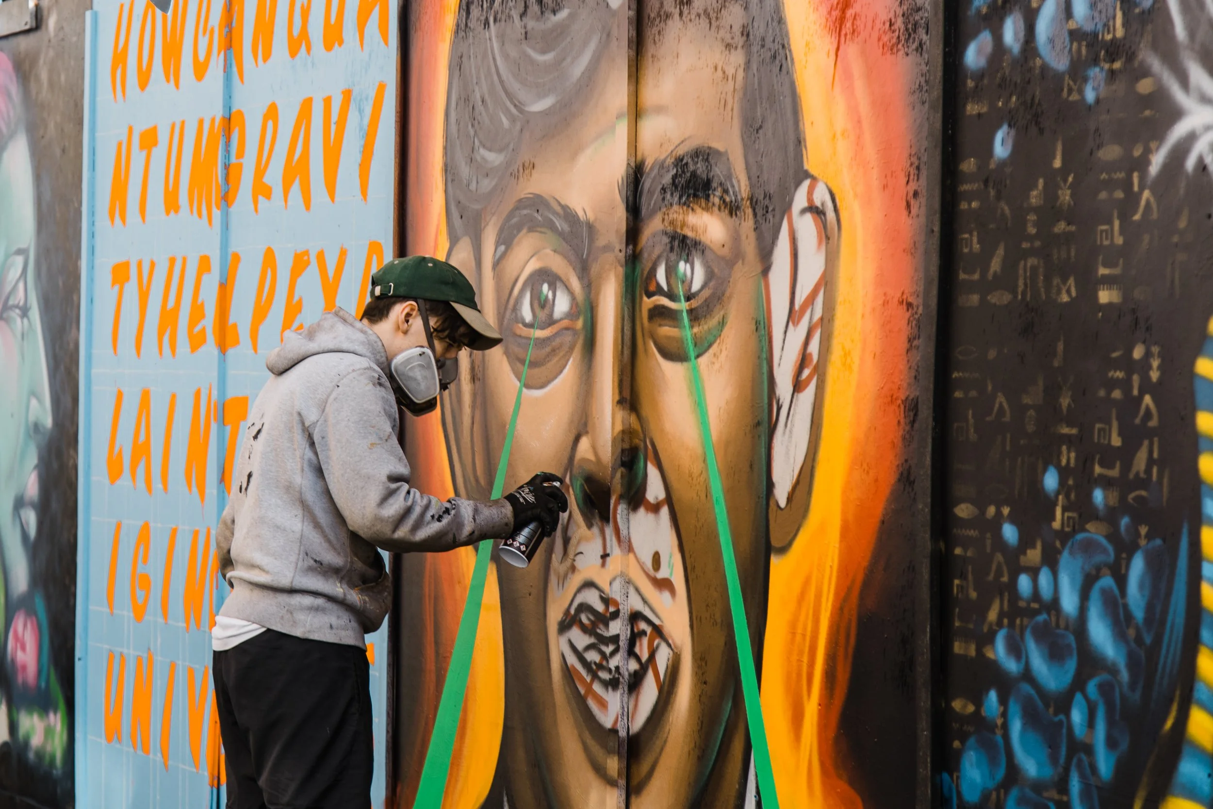 A person wearing a gray hoodie, black gloves, and a dark green cap spray paints graffiti art on a large wall. The mural includes a large eye and a face with colors blending in a spray paint style. The wall has orange text and various colorful artistic designs.