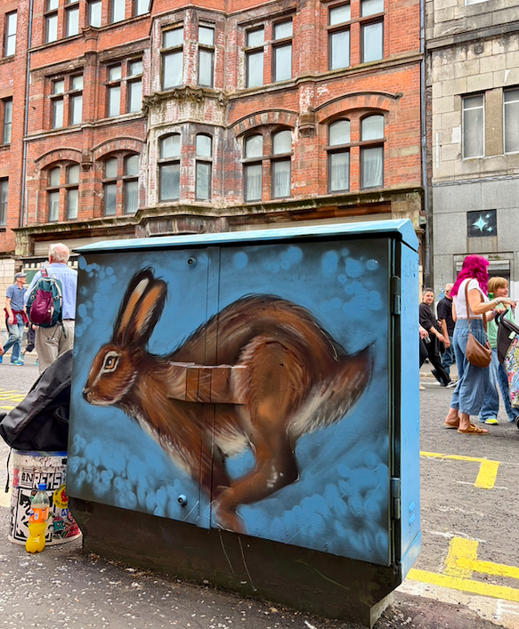 Street scene with a colorful mural of a running brown hare painted on an electrical box, with pedestrians walking in the background on a city street.