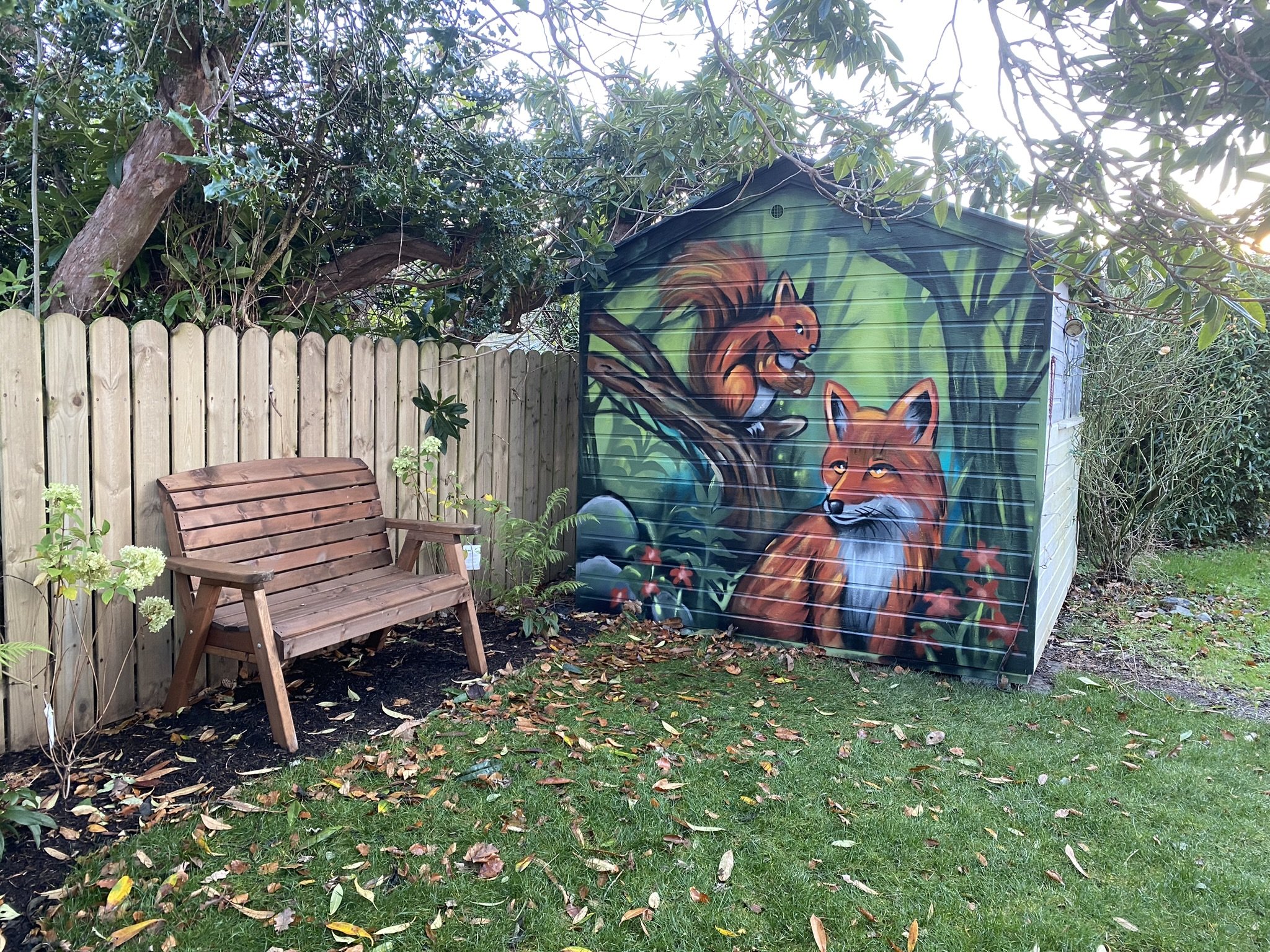 A backyard scene with a wooden fence, a wooden bench, a shed with graffiti art of a fox, and greenery including trees and plants. Leaves are scattered on the grass.